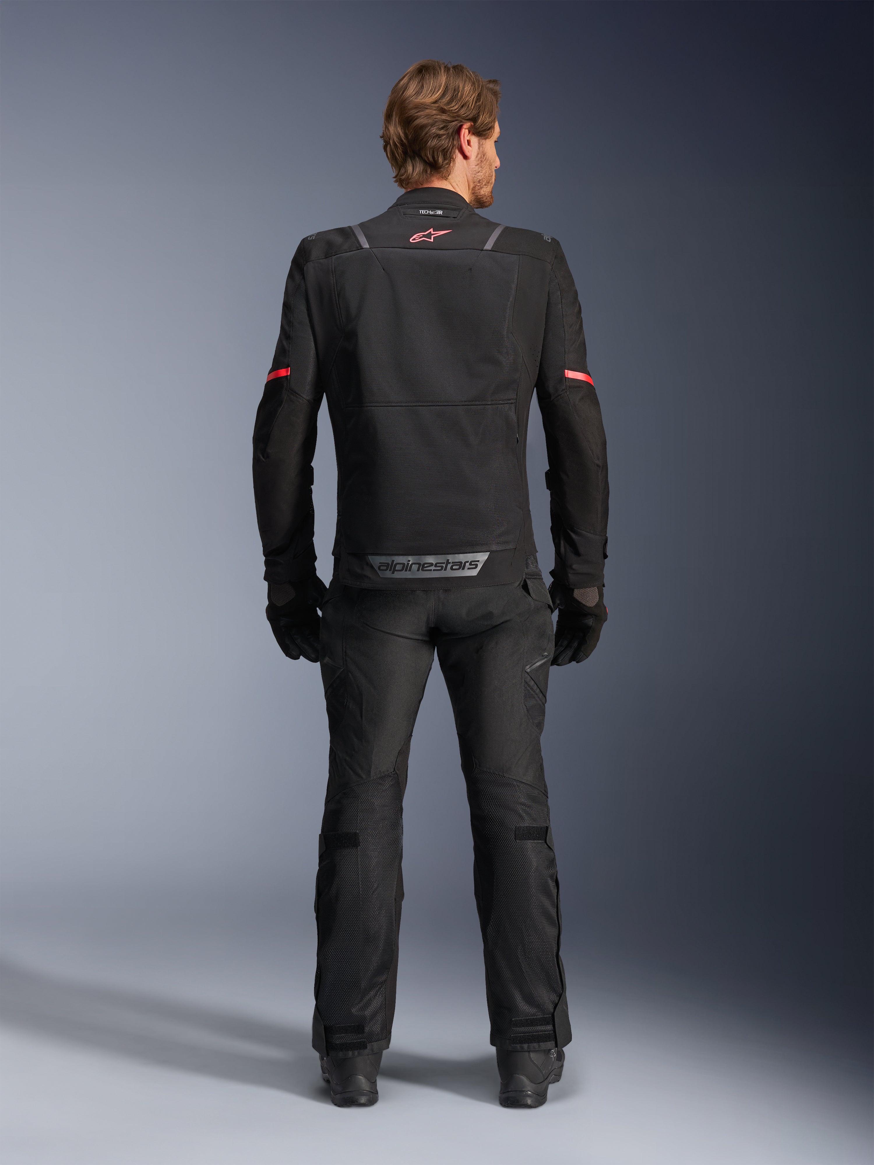 Alpinestars ST-2 Air jakke, sort, set bagfra af en kører iført motorcykeljakken og matchende sorte bukser, med mesh-ventilationspaneler, rødt logo øverst på ryggen og grå Alpinestars-branding på den nederste rygkant