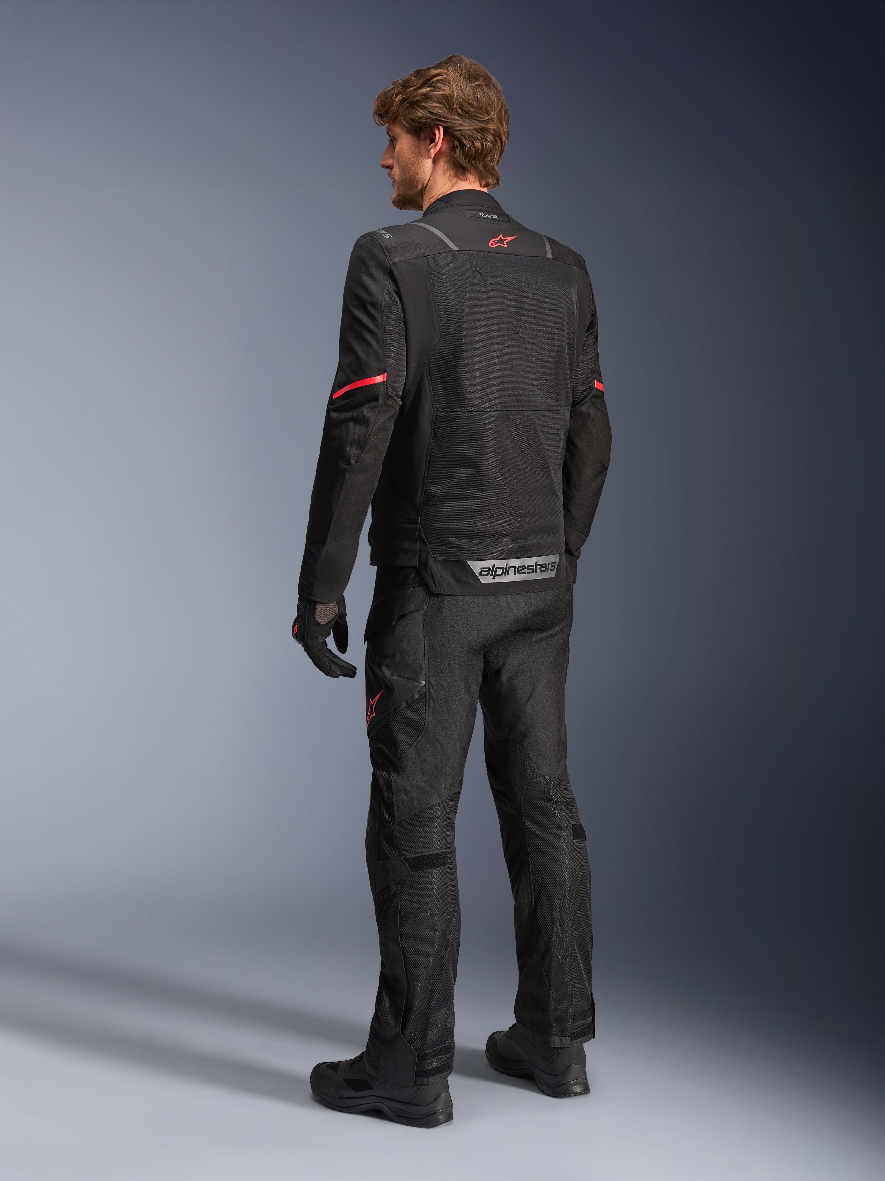 Alpinestars ST-2 Air Jakke, sort motorcykeljakke, set bagfra på model med et rødt stjernelogo øverst på ryggen og reflekterende Alpinestars-branding ved taljen, kombineret med matchende sorte tekniske kørebukser og handsker.