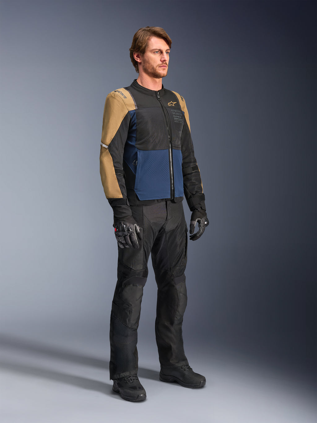 Mandlig motorcyklist iført Alpinestars ST-2 Air-jakke i Deep Blue, Dark Khaki og sort, med store mesh-paneler og laserperforeringer, kombineret med sorte motorcykelbukser, handsker og støvler for et komplet sommer-touring-look.