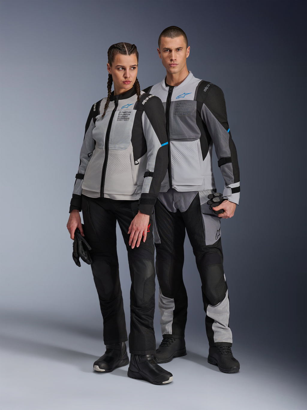 Mand og kvinde iført Alpinestars Stella ST-2 Air-jakker og matchende tekniske bukser, Ice Gray og sort farvekombination med blå detaljer, mesh-ventilation, 'ALPINESTARS EXPLORATION EQUIPMENT'-branding, studie-parbillede