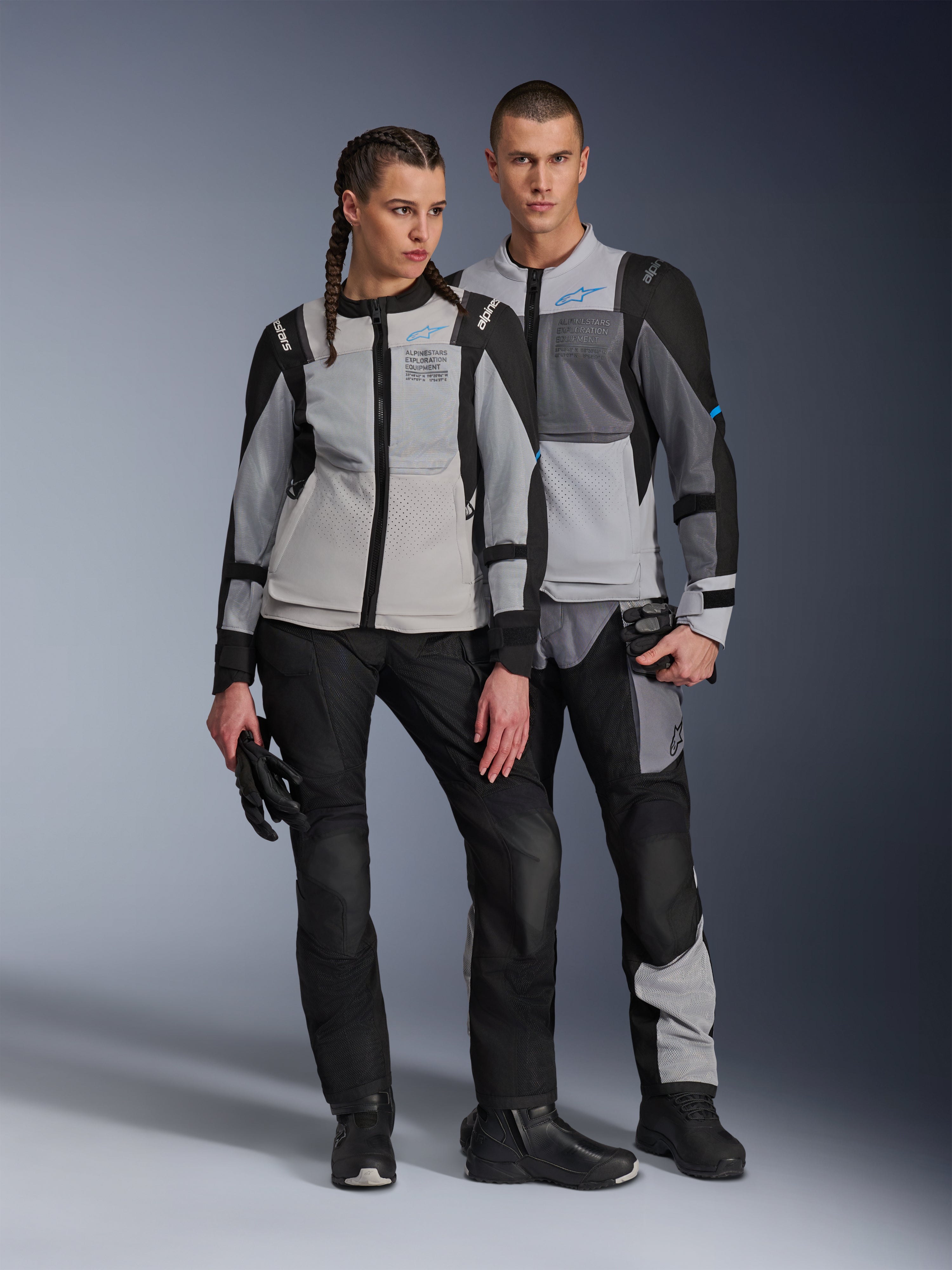 Mandlige og kvindelige modeller iført Alpinestars ST-2 Air-jakker i Ice Gray, Dark Gray og Black, teknisk motorcykeludstyr til sommertouring med store mesh-paneler og exploration equipment-branding, kombineret med sorte kørebukser, handsker og støvler