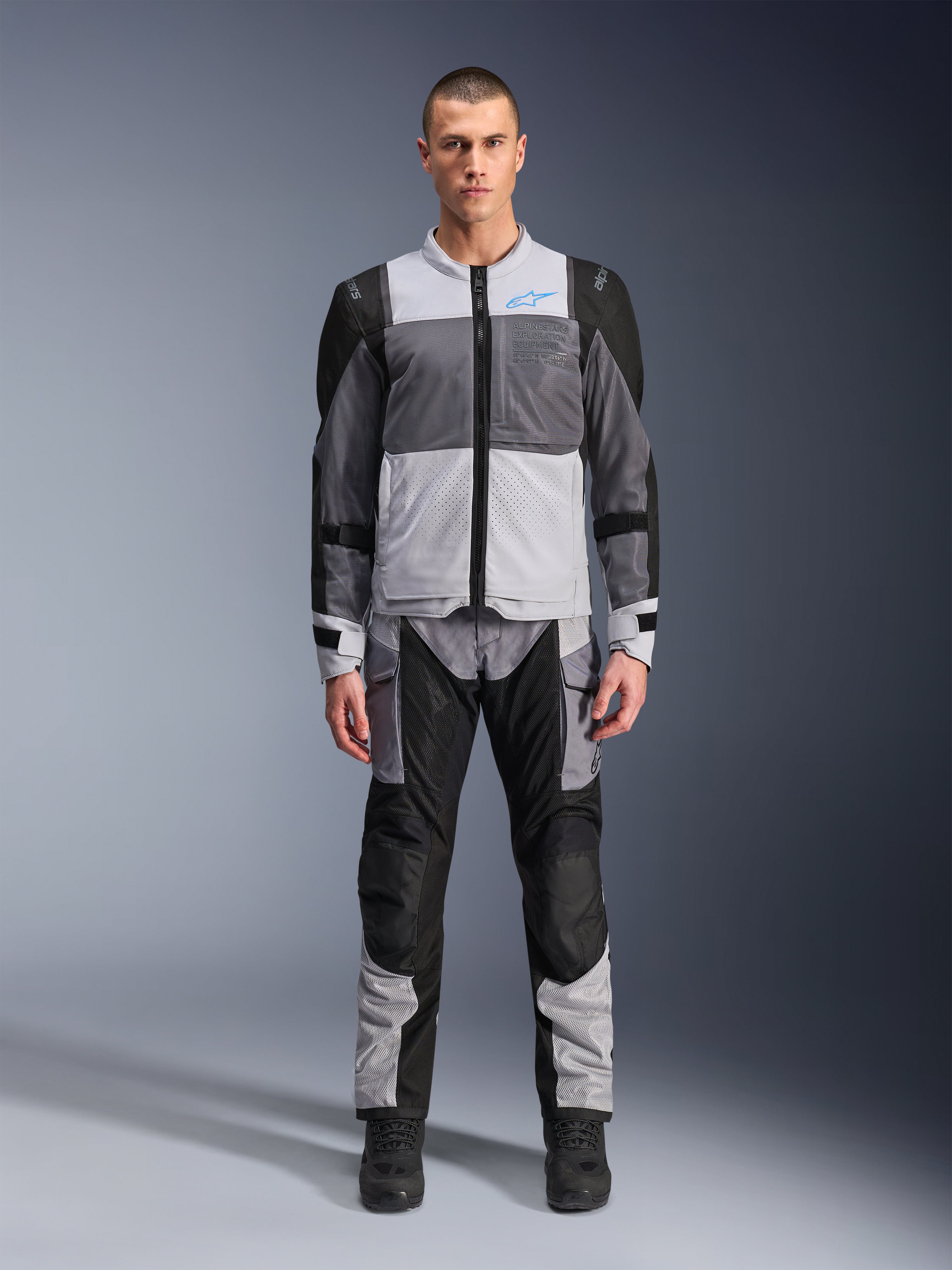 Alpinestars ST-2 Air Jakke, motorcykel touring-jakke, Ice Gray, Dark Gray og sort, båret af en model med matchende adventure-bukser, med store mesh-paneler, blåt Alpinestars-logo på brystet og Alpinestars Exploration Equipment-branding