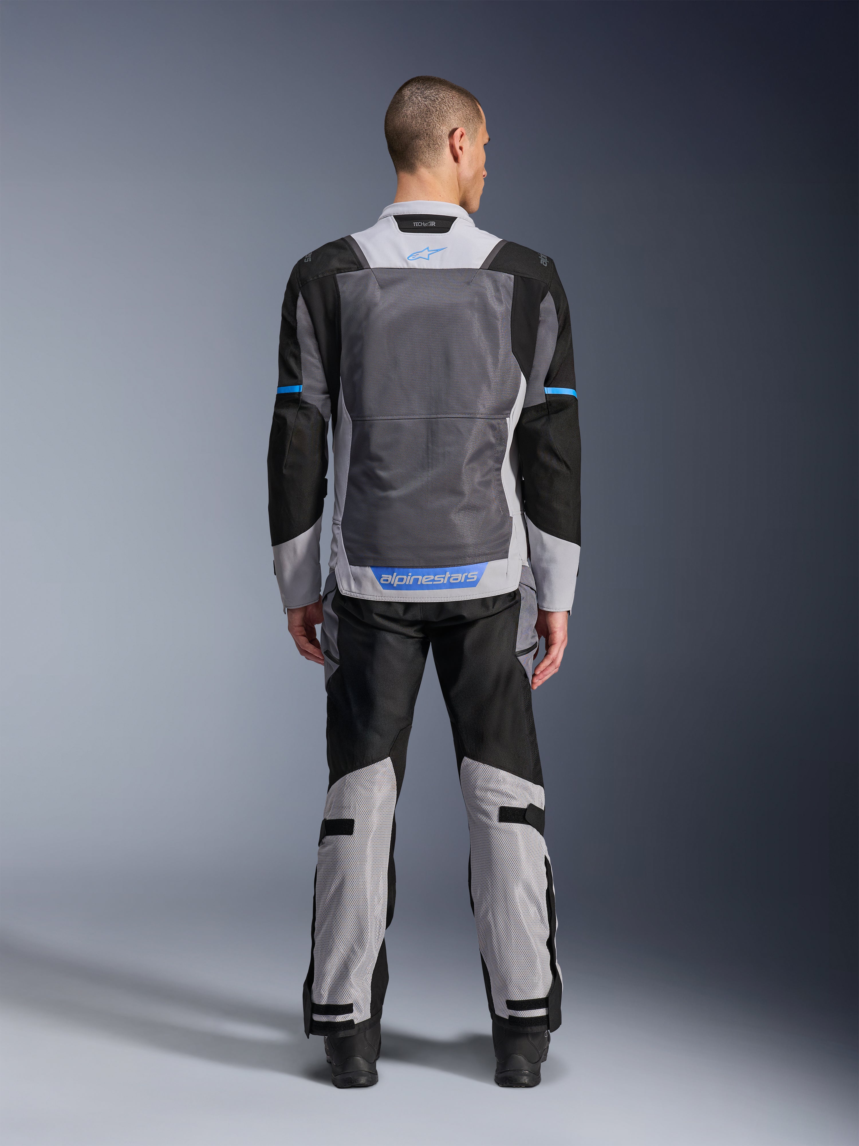 Alpinestars ST-2 Air-jakke, isgrå, mørkegrå og sort, set bagfra på model, med store mesh-ventilationspaneler, blå detaljer og Alpinestars-logo, kombineret med matchende motorcykelbukser til adventure touring.