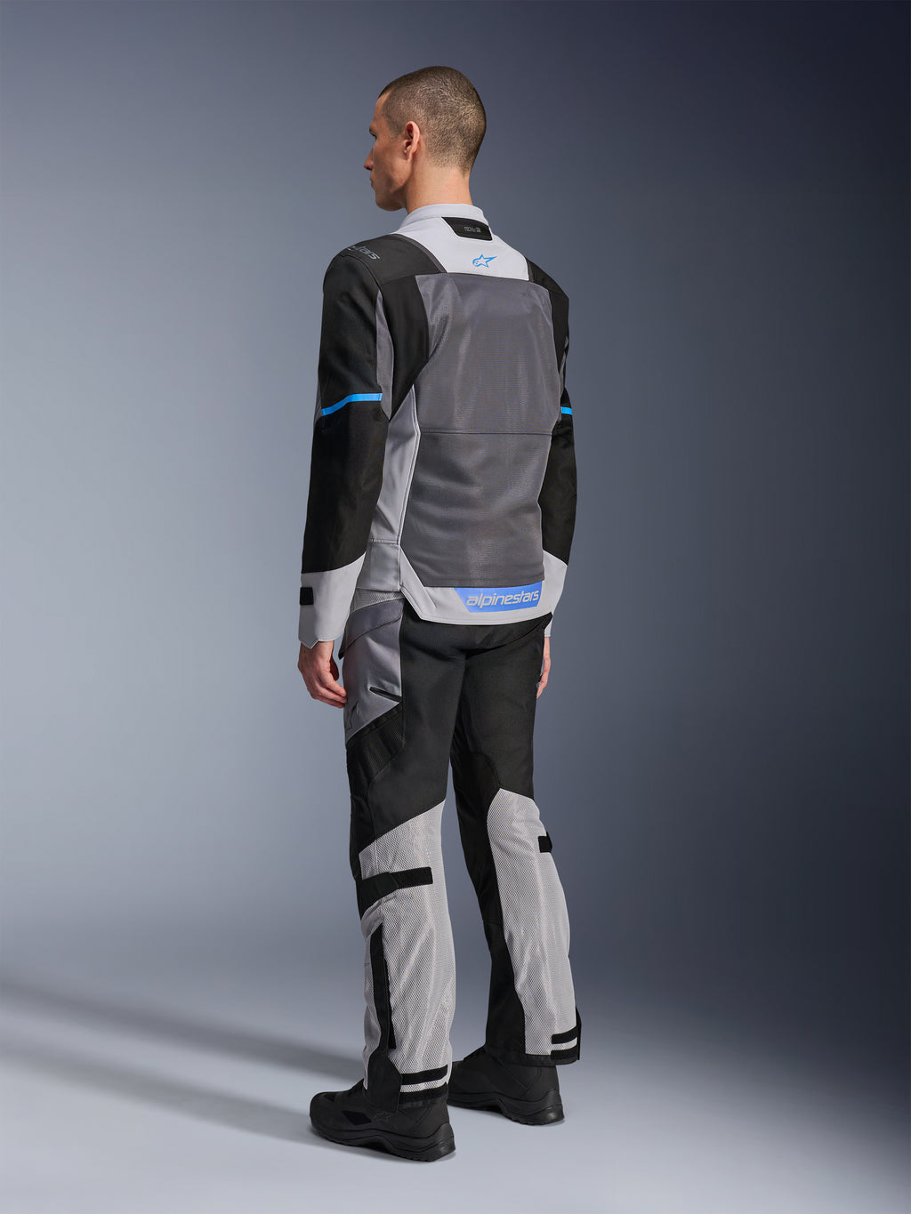 Alpinestars ST-2 Air-jakke, mandlig model set bagfra i Ice Gray, Dark Gray og sort, med stort gråt mesh-rygpanel, blå logo-detaljer og matchende motorcykelbukser til sommer-touring.