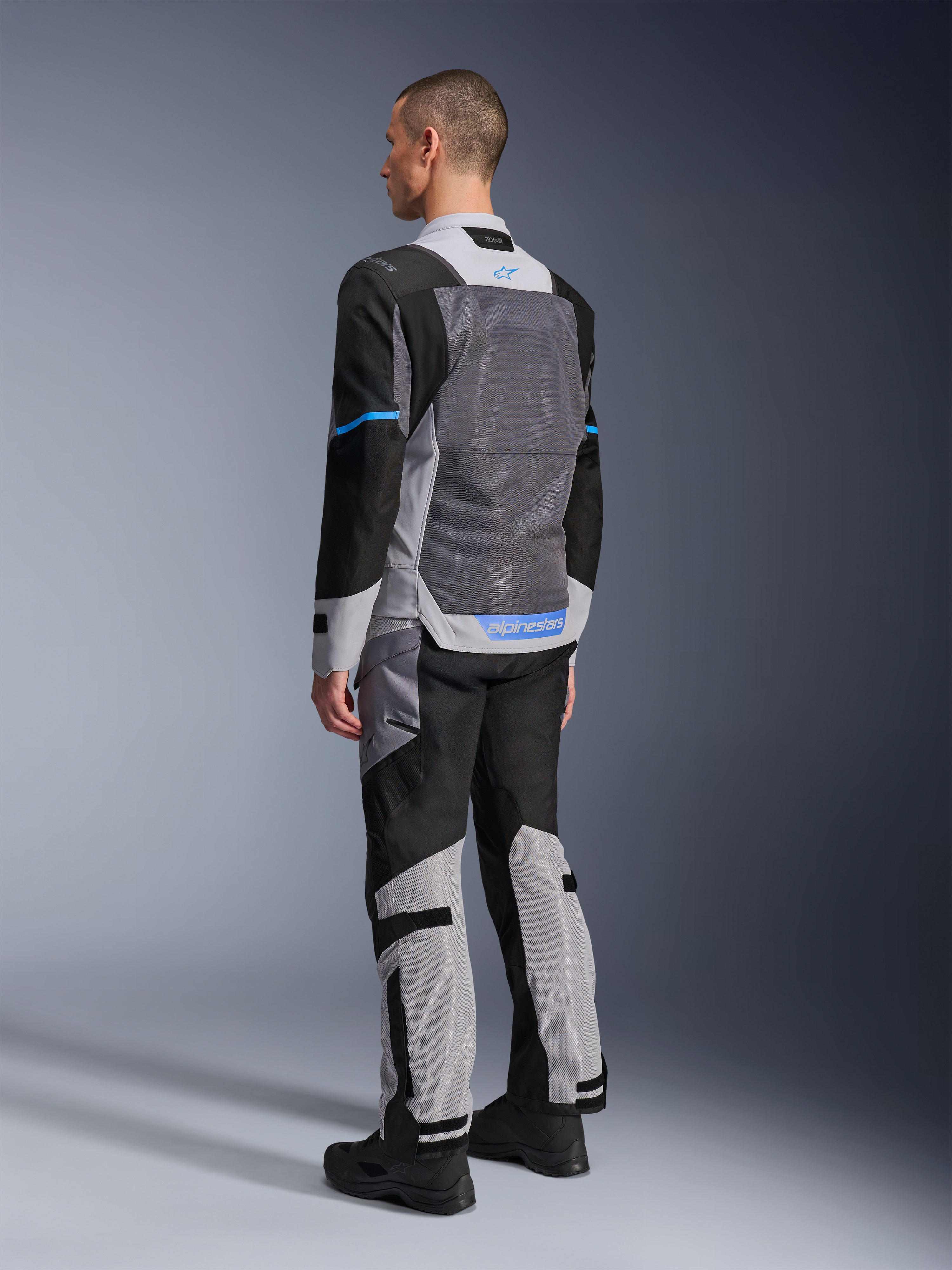 Alpinestars ST-2 Air-jakke, mandlig model set bagfra i Ice Gray, Dark Gray og sort, med stort gråt mesh-rygpanel, blå logo-detaljer og matchende motorcykelbukser til sommer-touring.