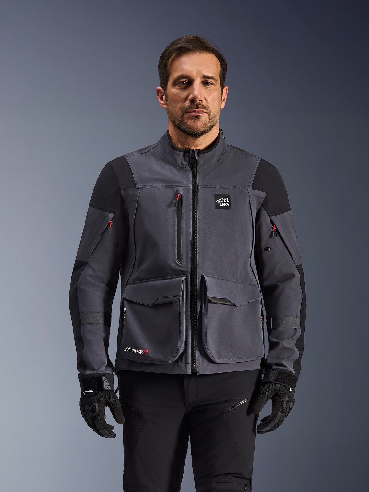 Alpinestars Maxdura Drystar®XF Laminated Armored Shell, mørkegrå og sort adventure-motorcykeljakke, set forfra på model, med store cargo-lommer, 'All Terra'-logo på brystet og ventilationslynlåse, kombineret med sorte kørehandsker og bukser.
