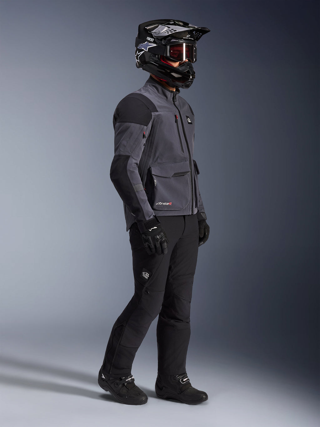 Motorcykelrytter iført Alpinestars Maxdura Drystar®XF Laminated Armored Shell-jakke i mørkegrå og sort, kombineret med sorte adventure-kørebukser, sort hjelm med goggles, sorte handsker og støvler, helkrops-studiebillede der fremhæver det tekniske dual-sport-udstyr.