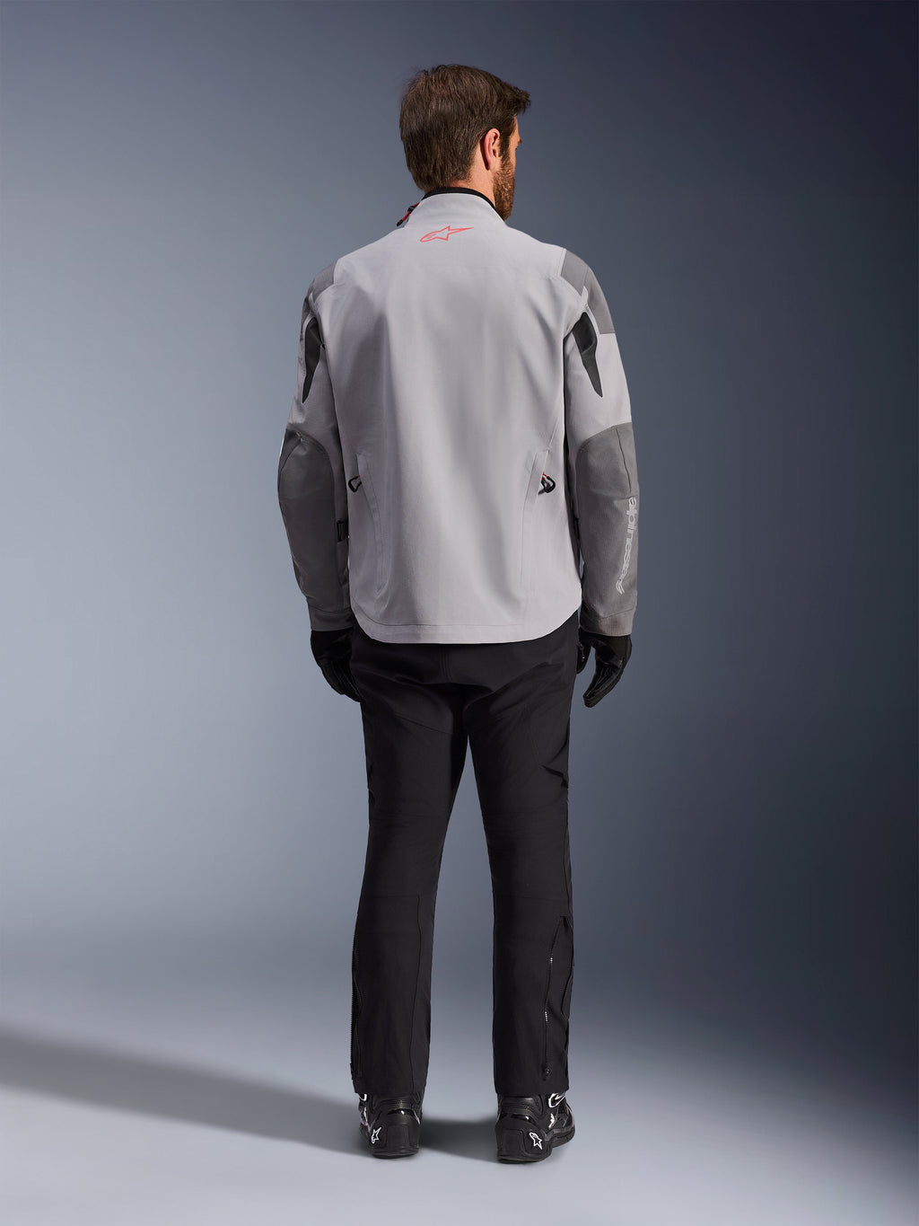 Alpinestars Maxdura Drystar®XF Laminated Armored Shell, mellemgrå og mørkegrå motorcykeljakke, set bagfra, med en lysegrå krop og mørkegrå paneler på skuldre og ærmer, lille rødt Alpinestars-stjernelogo på kraven bagpå og hvid branding på venstre underærme, kombineret med sorte kørebukser og støvler.