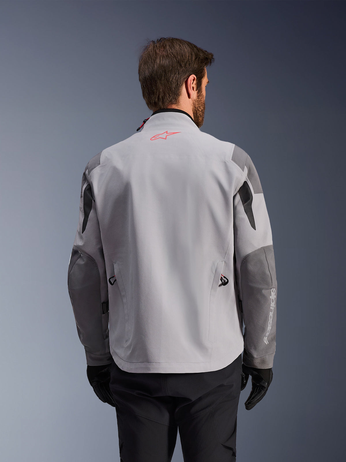 Alpinestars Maxdura Drystar®XF Laminated Armored Shell, adventure motorcykeljakke set bagfra, mellemgrå og mørkegrå, med rødt stjernelogo på kraven og hvidt lodret branding på ærmet, båret med sorte kørehandsker og bukser