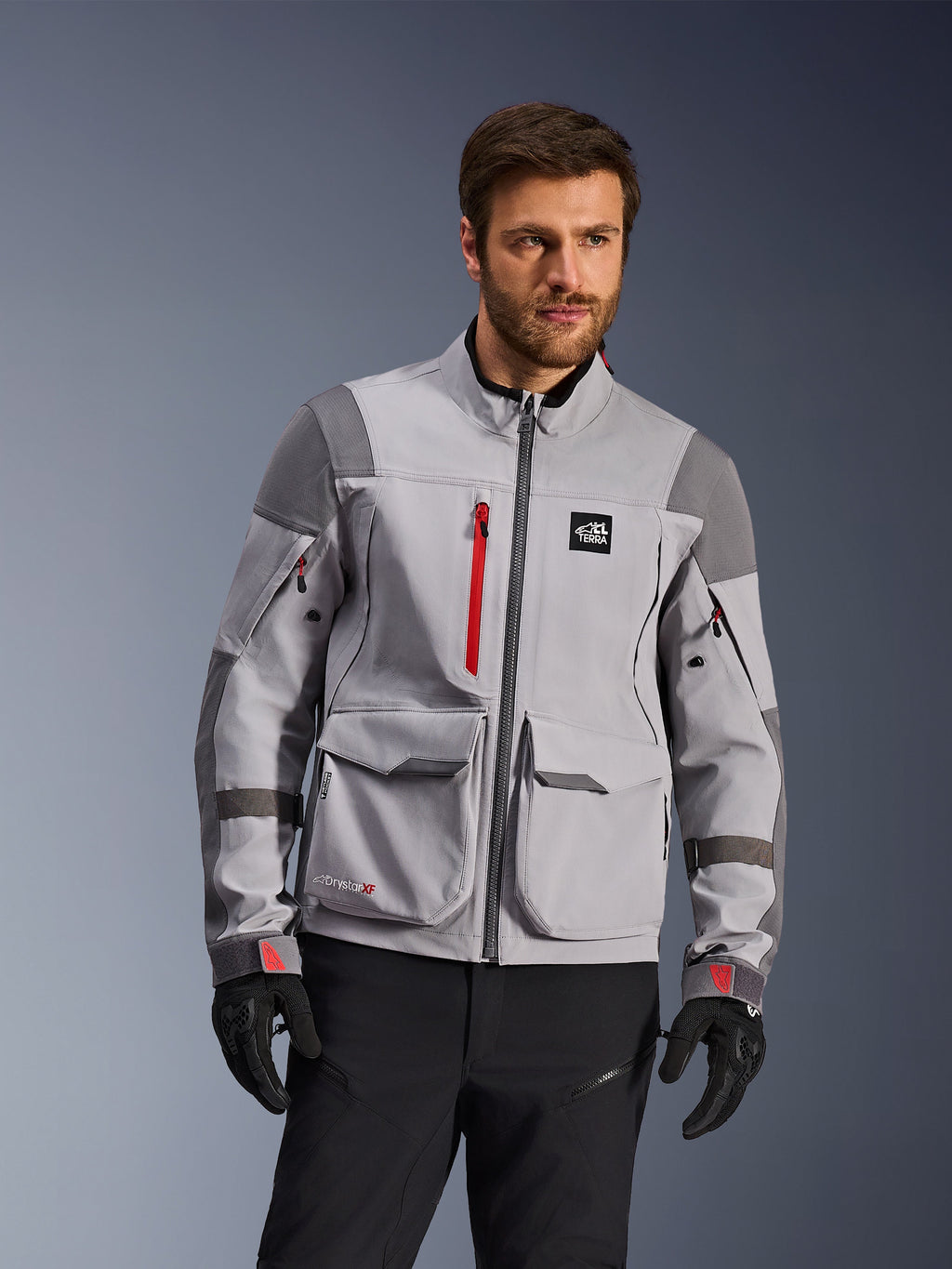 Alpinestars Maxdura Drystar®XF lamineret adventure motorcykeljakke, mellemgrå og mørkegrå med røde detaljer, bæres af en model med sorte handsker og bukser, med store cargo-lommer og vandtætte lynlåse, professionelt køreudstyr