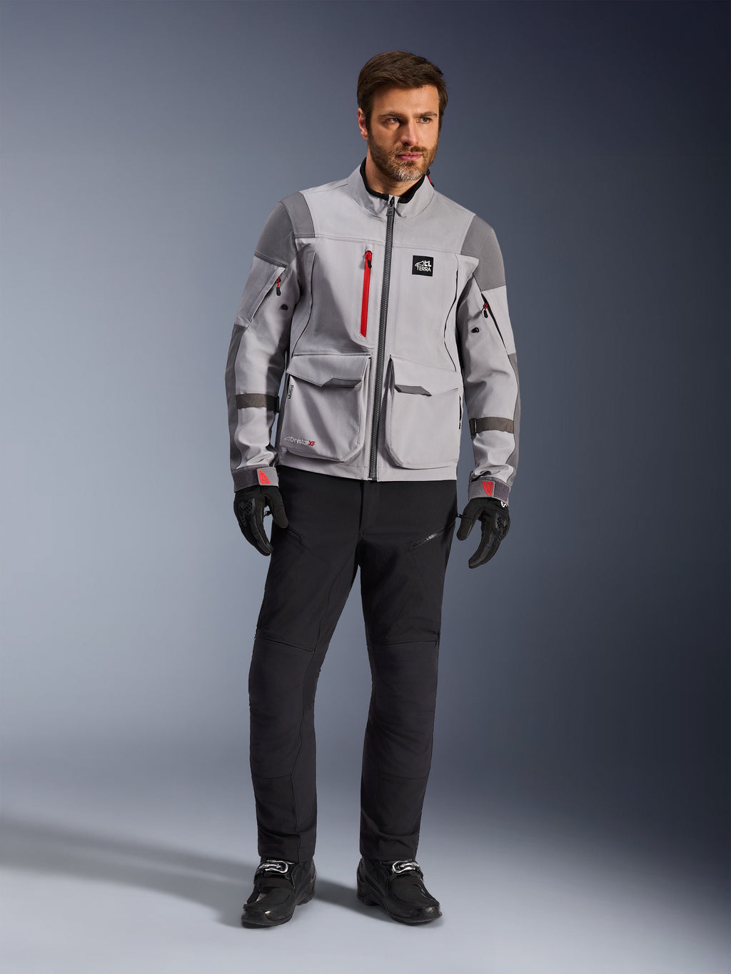Alpinestars Maxdura Drystar®XF Laminated Armored Shell, adventure motorcykeljakke, mellemgrå og mørkegrå med røde detaljer, båret med sorte kørebukser og handsker, set forfra med fokus på den tekniske konstruktion og store cargo-lommer.