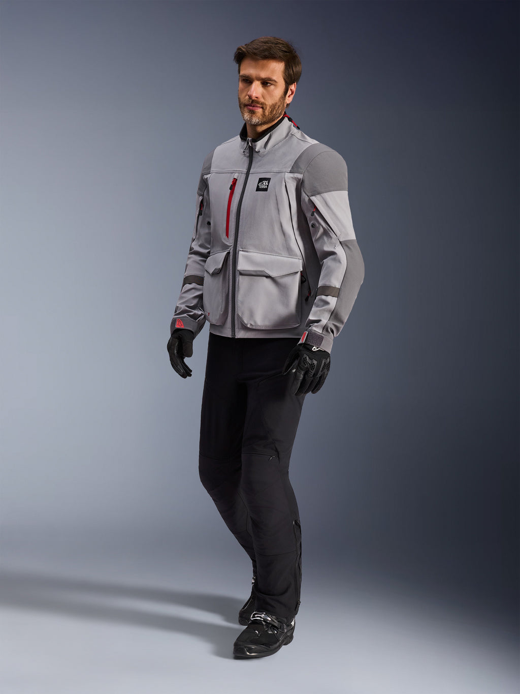 Alpinestars Maxdura Drystar®XF Lamineret Armored Shell, adventure-motorcykeljakke, mellemgrå og mørkegrå, båret af en model med sorte kørebukser og handsker, med røde lynlåsdetaljer og cargo-lommer, professionelt teknisk udstyr til al slags vejr til adventure touring