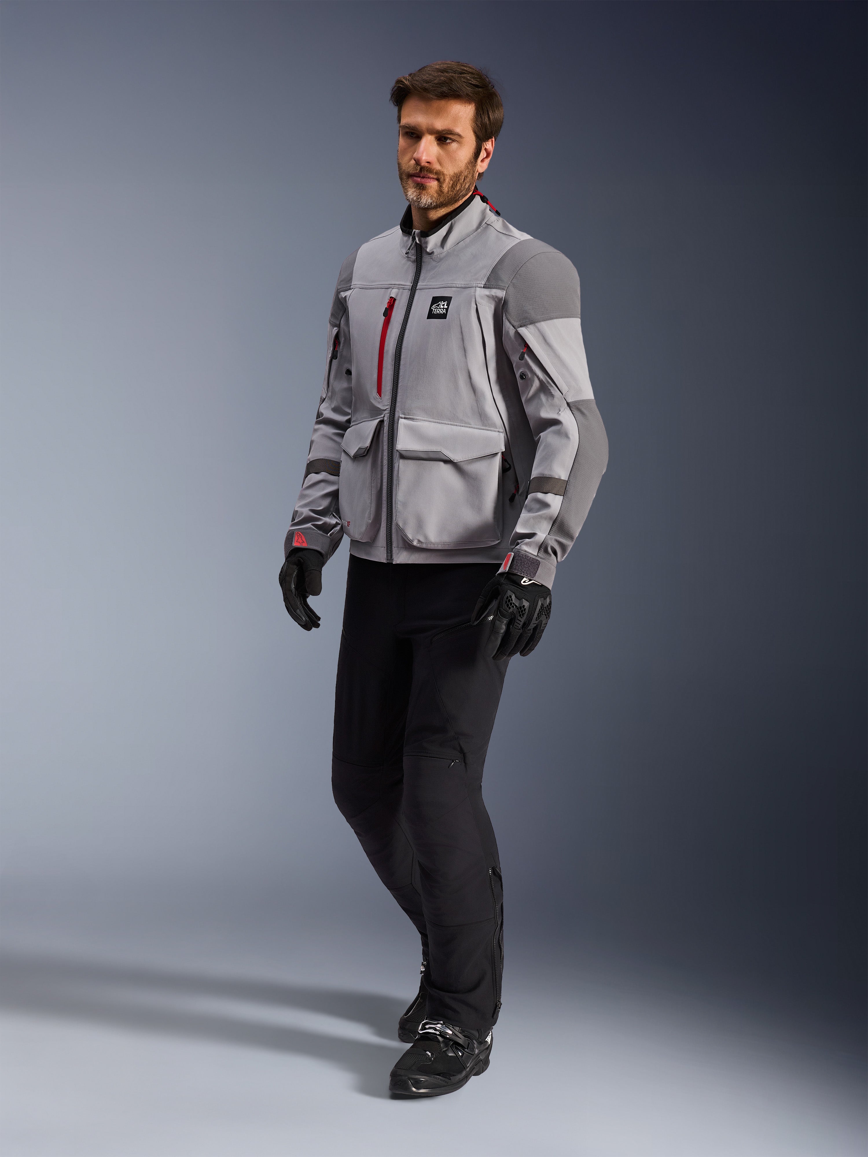 Alpinestars Maxdura Drystar®XF Lamineret Armored Shell, adventure-motorcykeljakke, mellemgrå og mørkegrå, båret af en model med sorte kørebukser og handsker, med røde lynlåsdetaljer og cargo-lommer, professionelt teknisk udstyr til al slags vejr til adventure touring