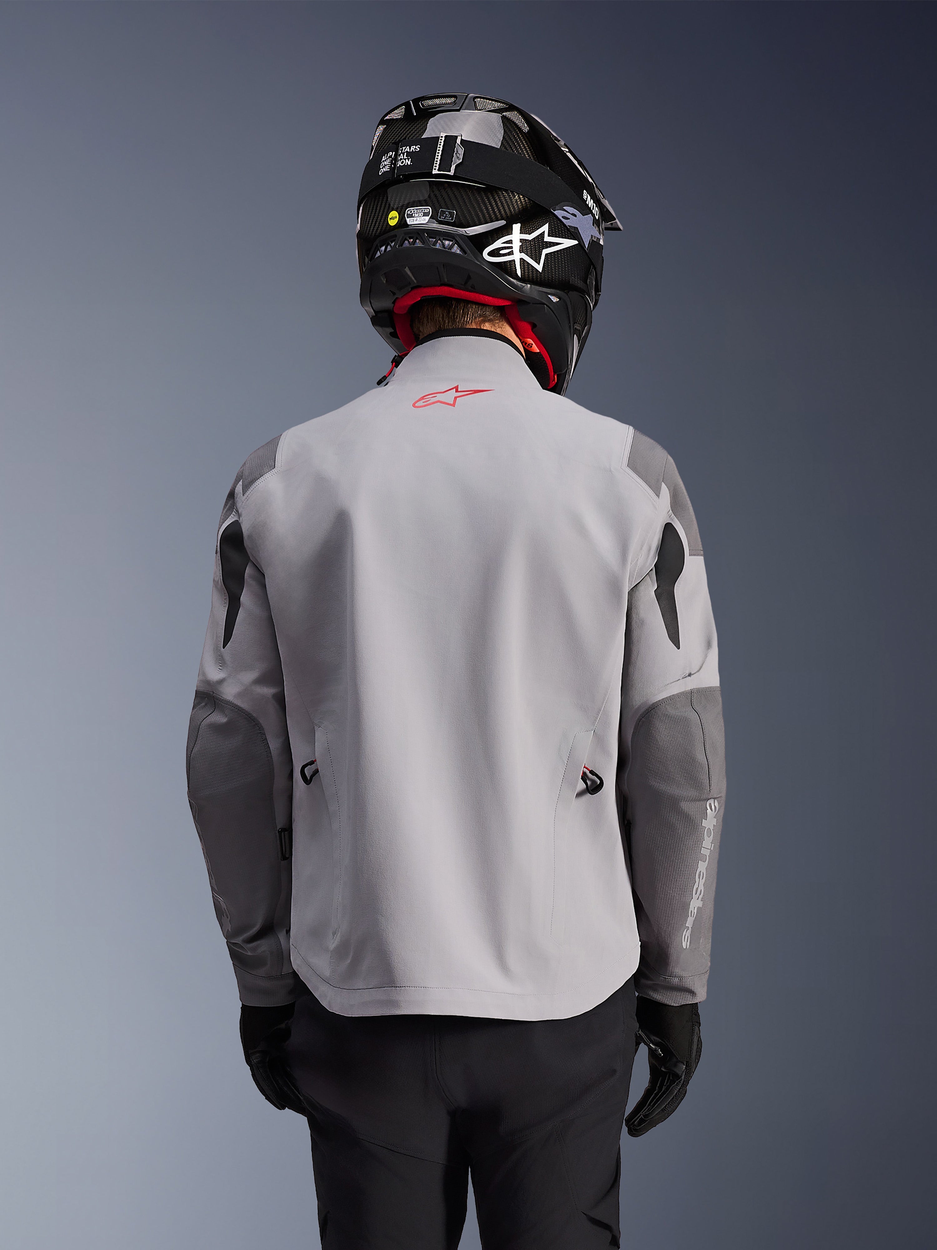Alpinestars Maxdura Drystar®XF Laminated Armored Shell, adventure motorcykeljakke, mellemgrå og mørkegrå, set bagfra med lysegrå krop og mørkere grå forstærkede paneler på skuldre og albuer, rødt Alpinestars-logo på øverste del af ryggen, båret med sort hjelm og sorte handsker.