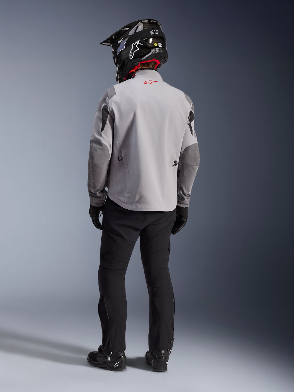 Set bagfra af en motorcykelrytter iført Alpinestars Maxdura Drystar®XF Laminated Armored Shell-jakken i Mid Gray og Dark Gray, med et rødt stjernelogo på kraven, kombineret med sorte adventure-bukser, en sort hjelm med carbon-mønster og sorte støvler.