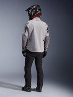 Set bagfra af en motorcykelrytter iført Alpinestars Maxdura Drystar®XF Laminated Armored Shell-jakken i Mid Gray og Dark Gray, med et rødt stjernelogo på kraven, kombineret med sorte adventure-bukser, en sort hjelm med carbon-mønster og sorte støvler.
