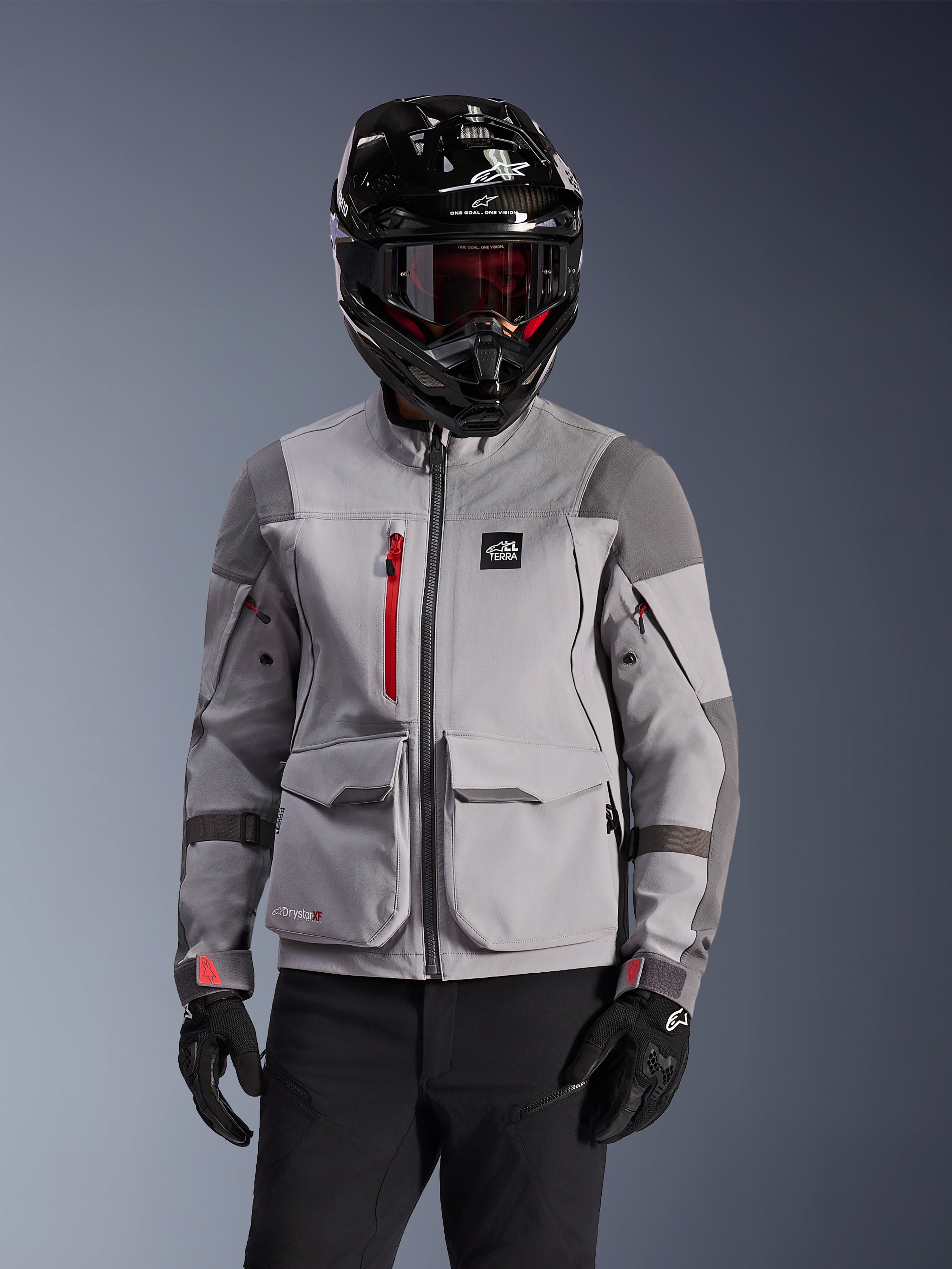 Alpinestars Maxdura Drystar®XF Lamineret Armored Shell, adventure-motorcykeljakke i mellemgrå og mørkegrå, båret med en sort hjelm med carbon-finish og rødtonede goggles, med front-cargolommer og en rød brystlynlås, teknisk design til adventure touring.