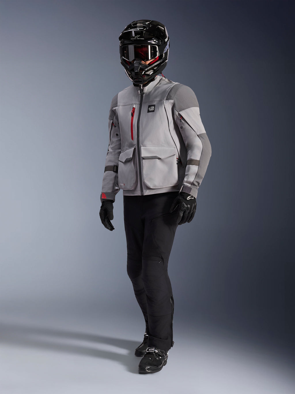Adventure-rytter iført Alpinestars Maxdura Drystar®XF lamineret motorcykeljakke i mellemgrå og mørkegrå, sort adventure-hjelm med crossbriller, sorte bukser og handsker, med røde lynlåsdetaljer og cargo-lommer, teknisk udstyr til adventure touring.