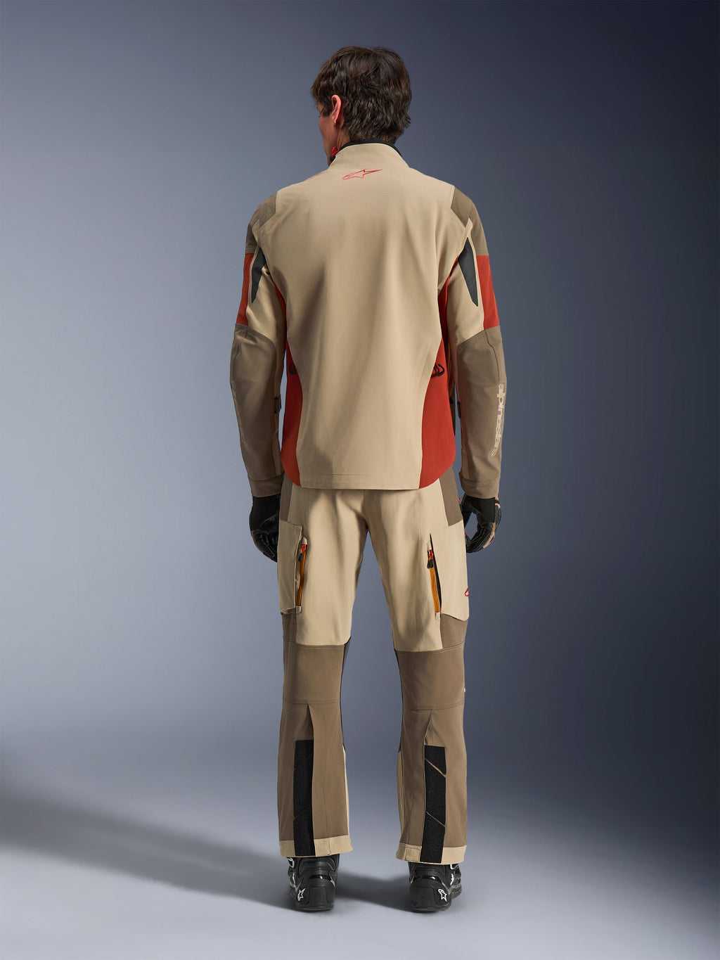 Set bagfra af en kører iført Alpinestars Maxdura Drystar®XF Laminated Armored Shell og matchende adventure-bukser i Aluminum Walnut Rust Brown, sandfarvet og brunt tekstil med orange detaljer, kombineret med sorte motorcykelstøvler og handsker, professionelt adventure touring-udstyr.