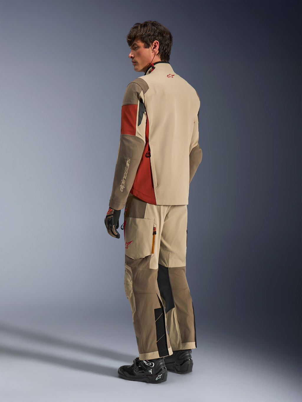 Mandlig model iført Alpinestars Maxdura Drystar®XF Laminated Armored Shell i Aluminum Walnut Rust Brown, matchende adventure touring-bukser, sorte handsker og sorte støvler, rygvisning der viser den tekniske tekstilkonstruktion og de forstærkede zoner.