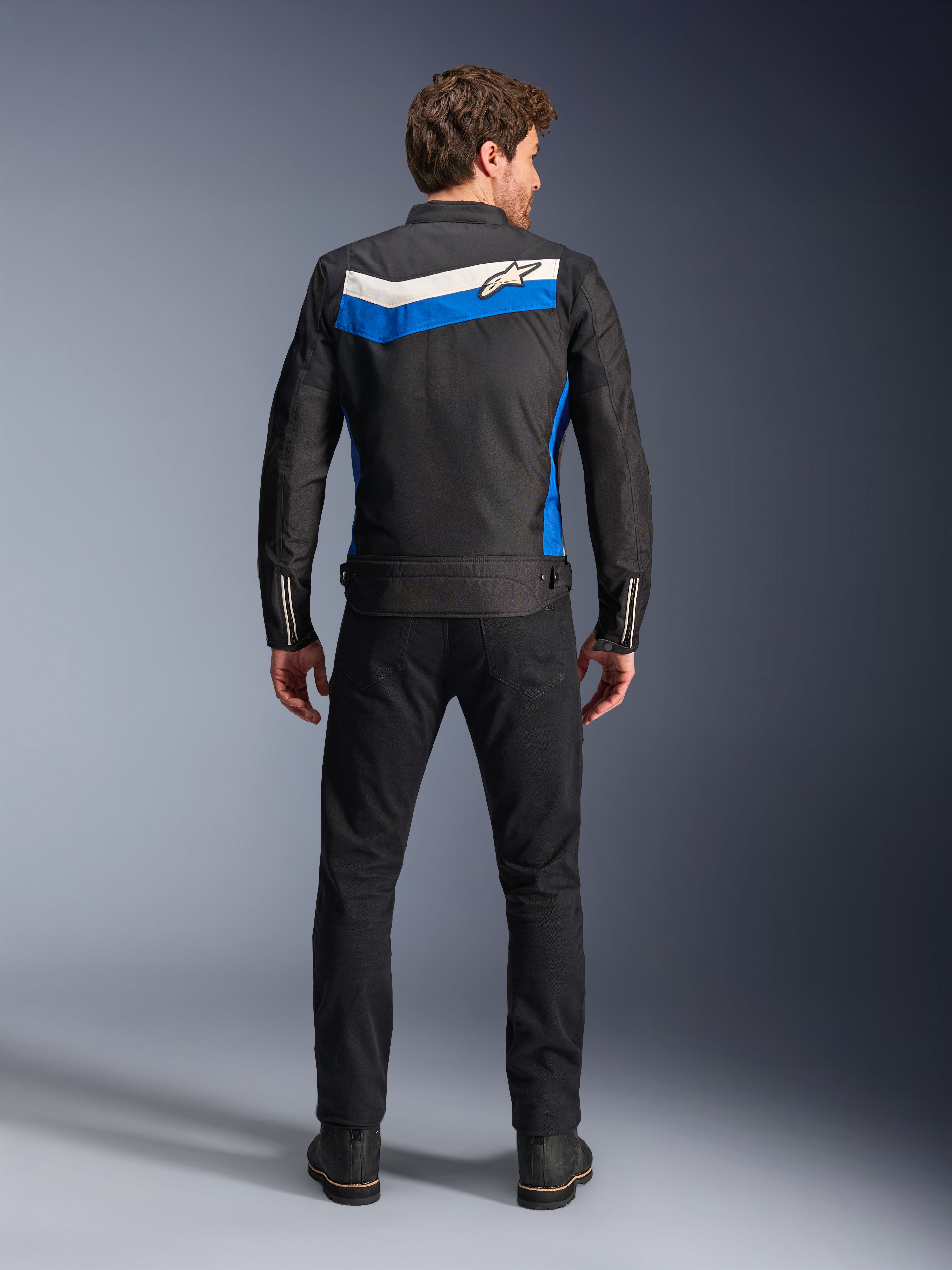 Alpinestars T-Dyno WR Jacket, textile motorcycle jacket, Black Blue Ecru colorway, full back view on model, featuring a blue and off-white chevron stripe with Alpinestars logo, paired with black pants and boots, vintage-inspired urban riding gear