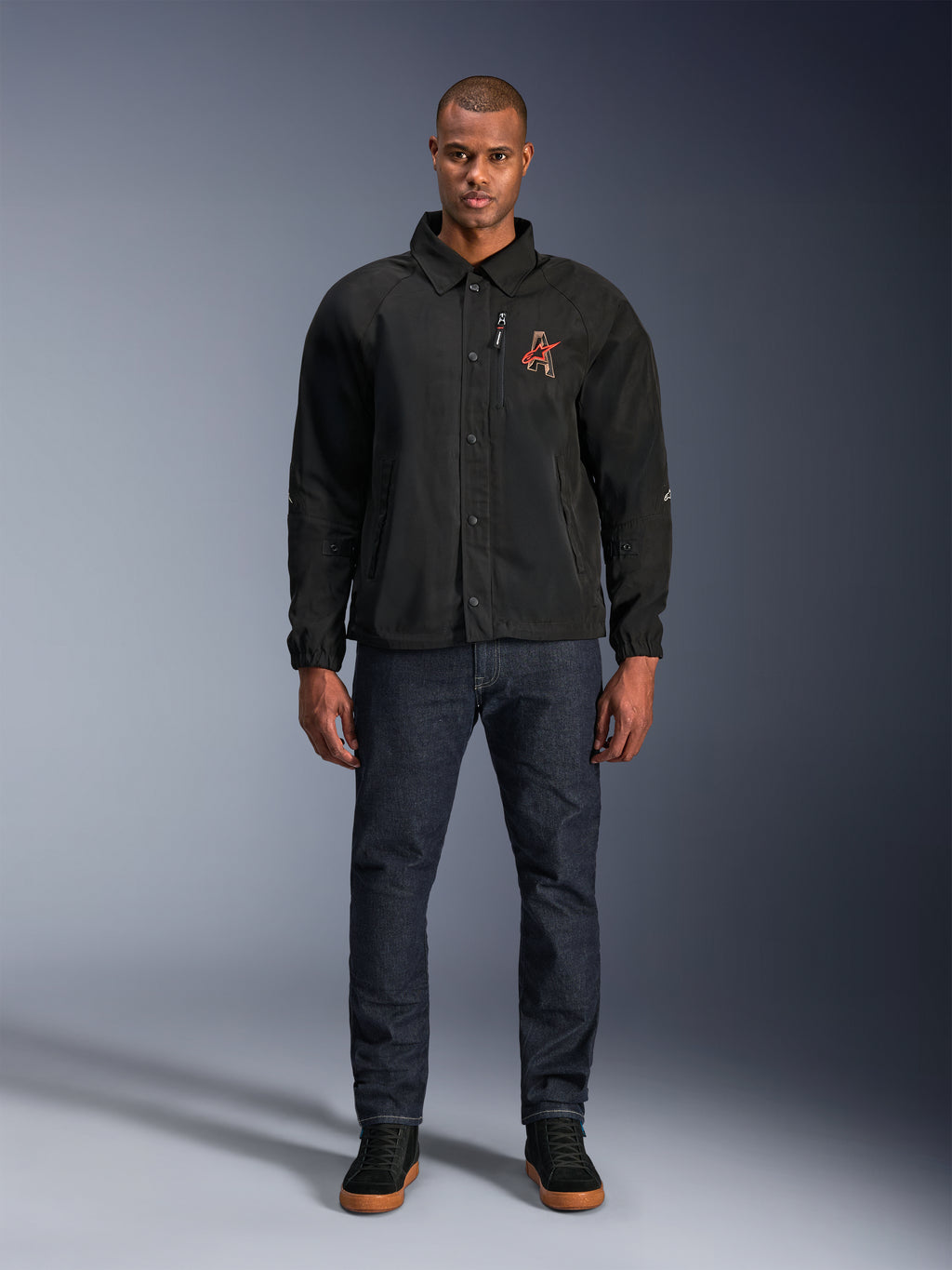 Alpinestars Revved WR-jakke i sort, båret af en model med mørkeblå jeans og sorte sneakers, set forfra, med trykknaplukning og et 3D-broderet logo på brystet, urbant moderne motorcykeludstyr
