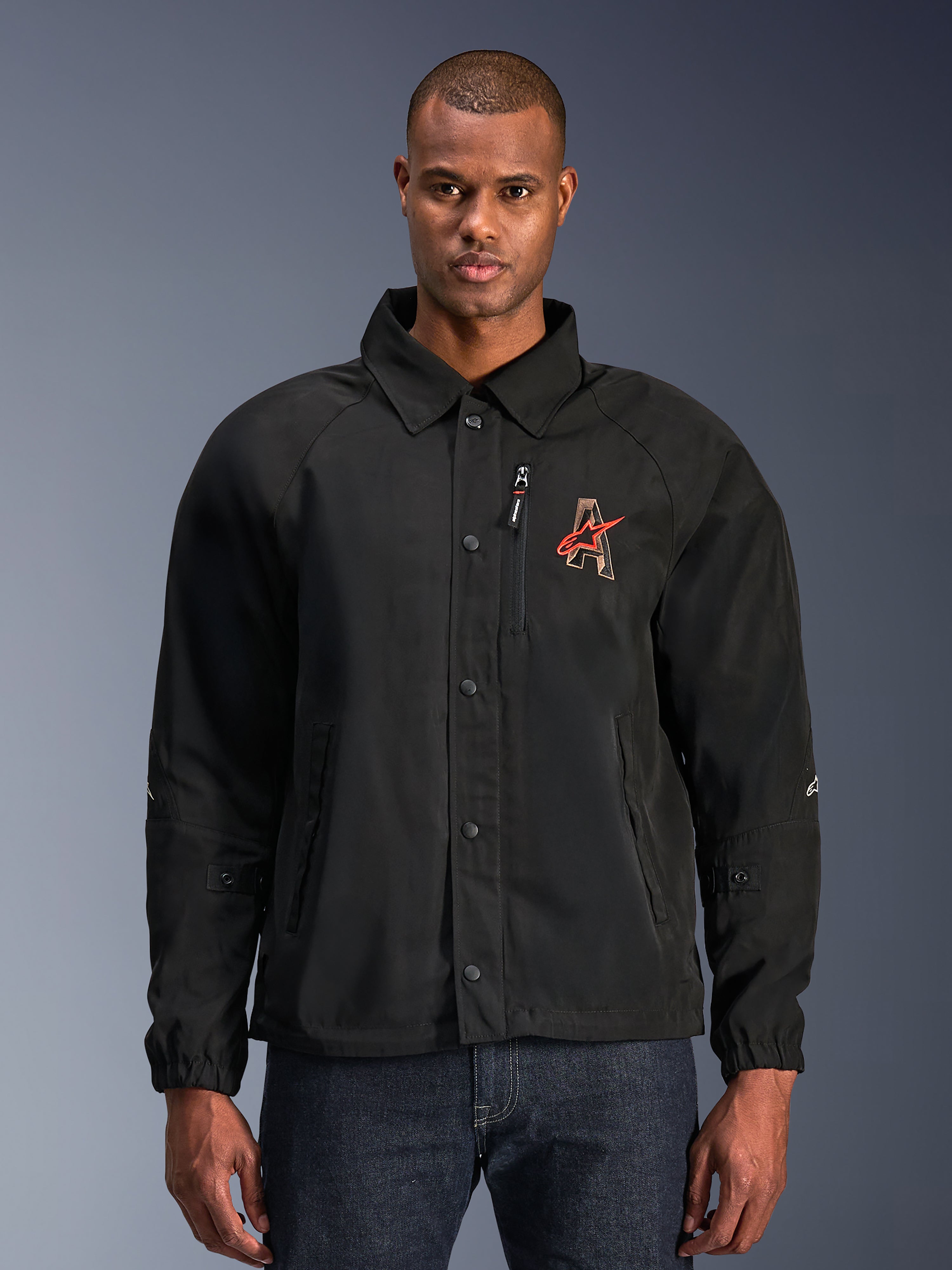 Alpinestars Revved WR-jakke, sort motorcykeljakke med skjortekrave og trykknapper foran, rødt broderet 'A'-logo på brystet, lodret brystlomme med lynlås, elastiske manchetter, urbant coach-jakke design
