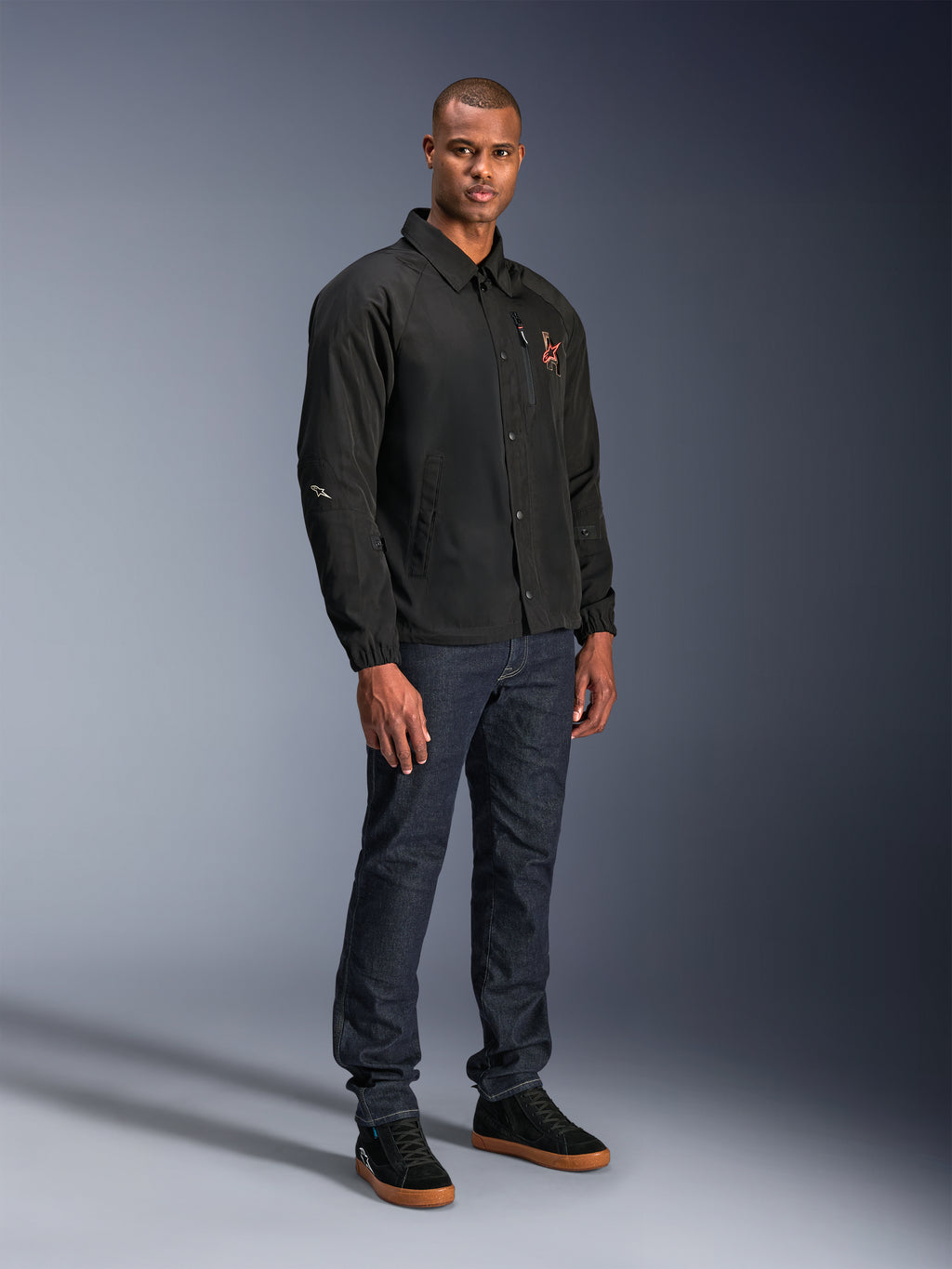 Alpinestars Revved WR-jakke i sort, urban motorcykeljakke i coach-stil, båret af en model med mørkeblå jeans og sorte sneakers, med trykknapper foran og broderet logo, minimalistisk og sporty design til landevejskørsel.