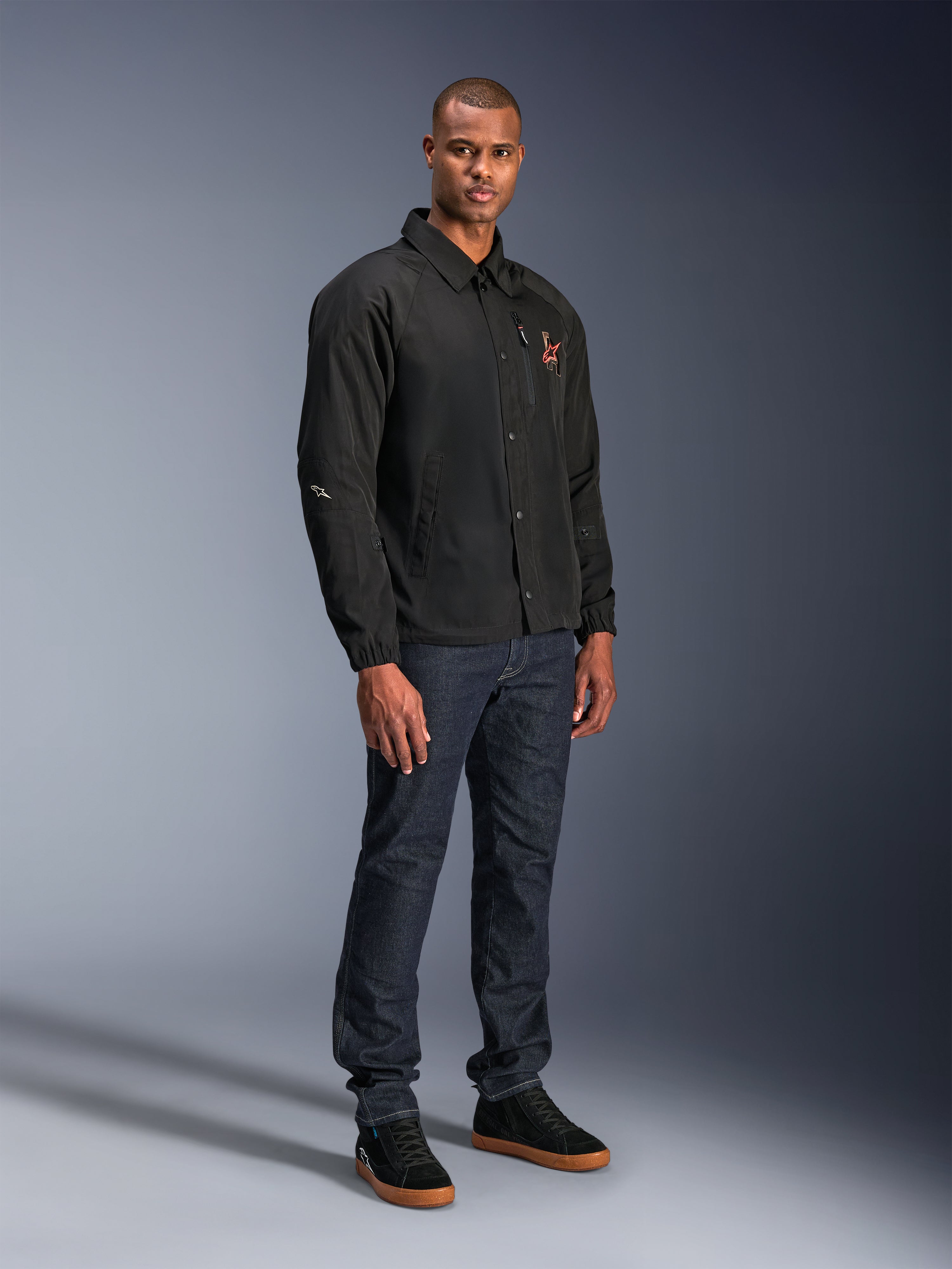 Alpinestars Revved WR-jakke i sort, urban motorcykeljakke i coach-stil, båret af en model med mørkeblå jeans og sorte sneakers, med trykknapper foran og broderet logo, minimalistisk og sporty design til landevejskørsel.