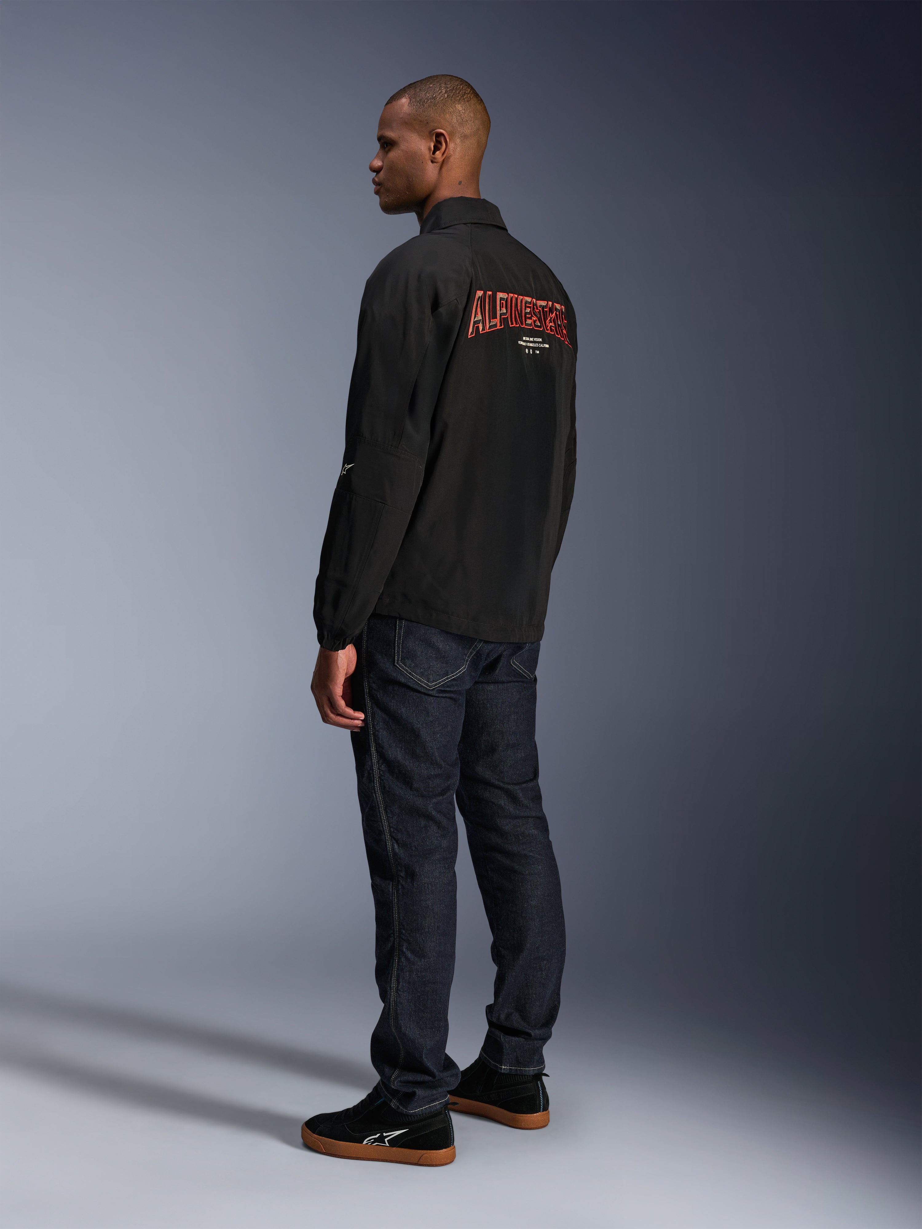 Alpinestars Revved WR-jakke, sort, med stort orange 3D-broderet Alpinestars-branding på ryggen, set bagfra i trekvart profil på en model iført mørkeblå jeans og sorte sneakers, urban moderne motorcykeljakke med coach-jakke-silhuet