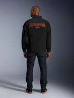 Alpinestars Revved WR Jakke, sort, set bagfra med stort rødt 'ALPINESTARS' broderi i 3D-effekt, urban coach-jakke, båret med mørke denimjeans og sorte sneakers, minimalistisk motorcykelbeklædning