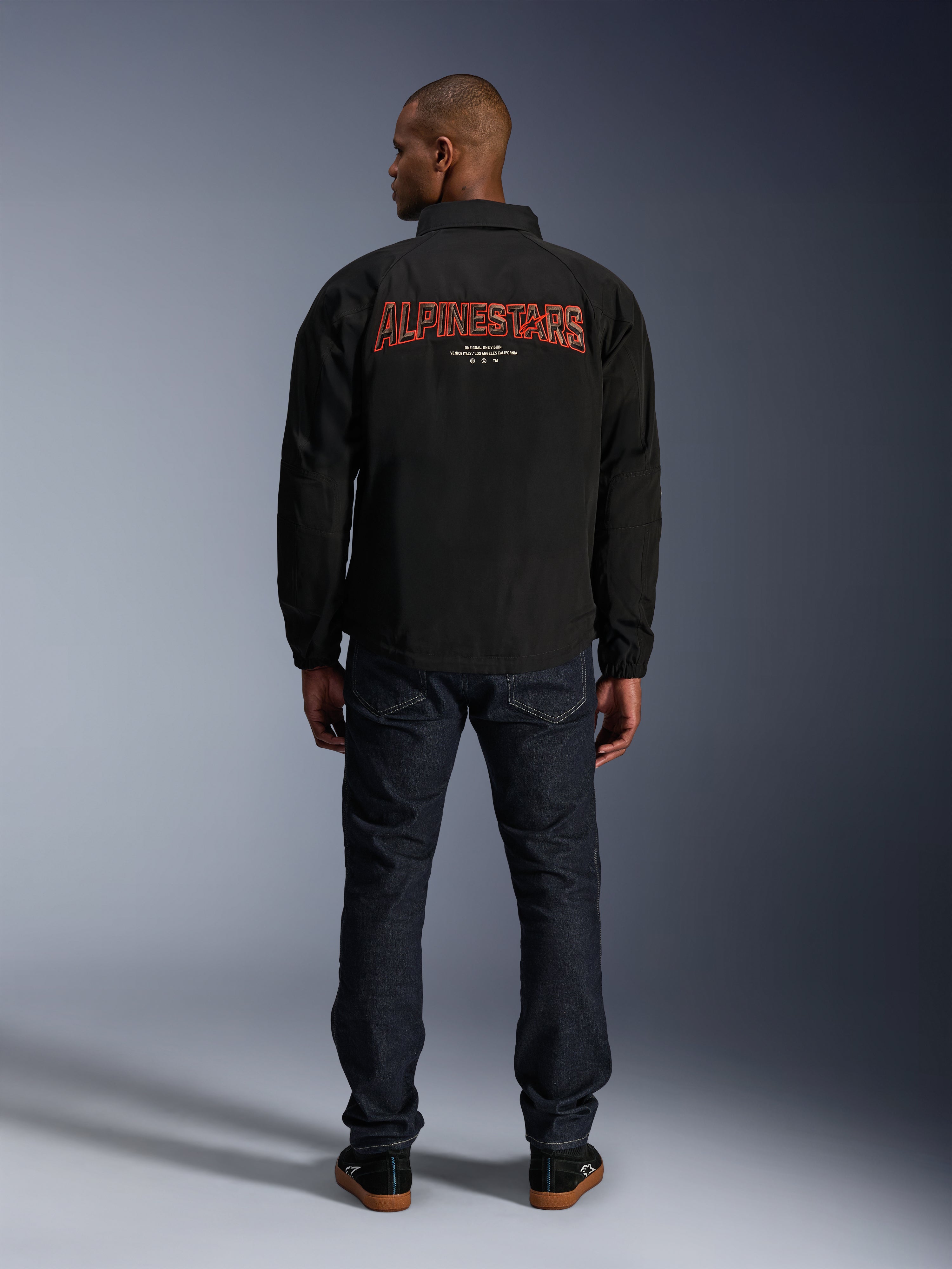 Alpinestars Revved WR Jakke, sort, set bagfra med stort rødt 'ALPINESTARS' broderi i 3D-effekt, urban coach-jakke, båret med mørke denimjeans og sorte sneakers, minimalistisk motorcykelbeklædning