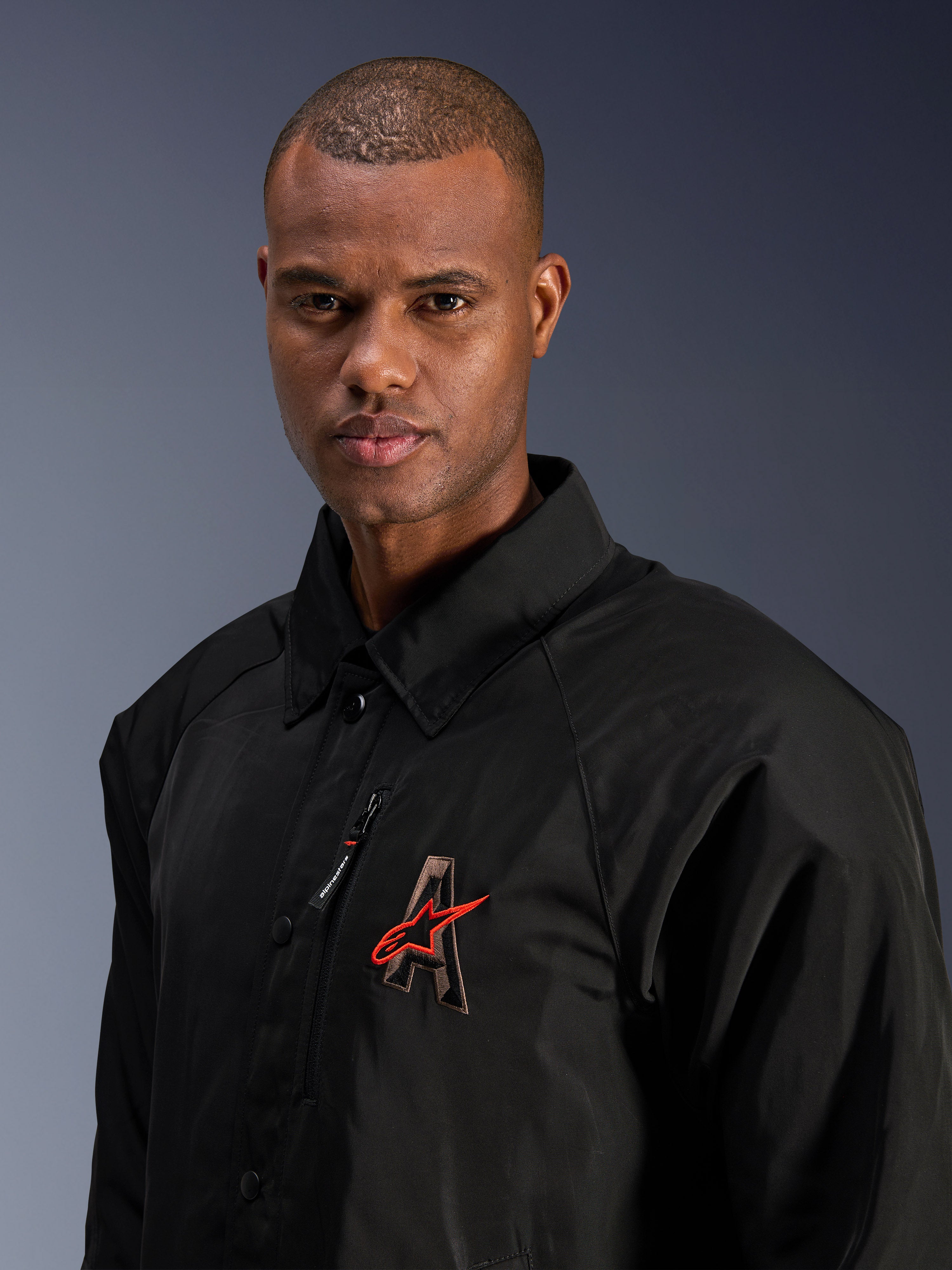 Alpinestars Revved WR Jakke, sort, nærbillede af en mand iført den vandafvisende motorcykeljakke i satin, med 3D-broderet 'A'-logo med rød Alpinestars-stjerne, brystlomme med lynlås og design med krave til moderne bykørsel.