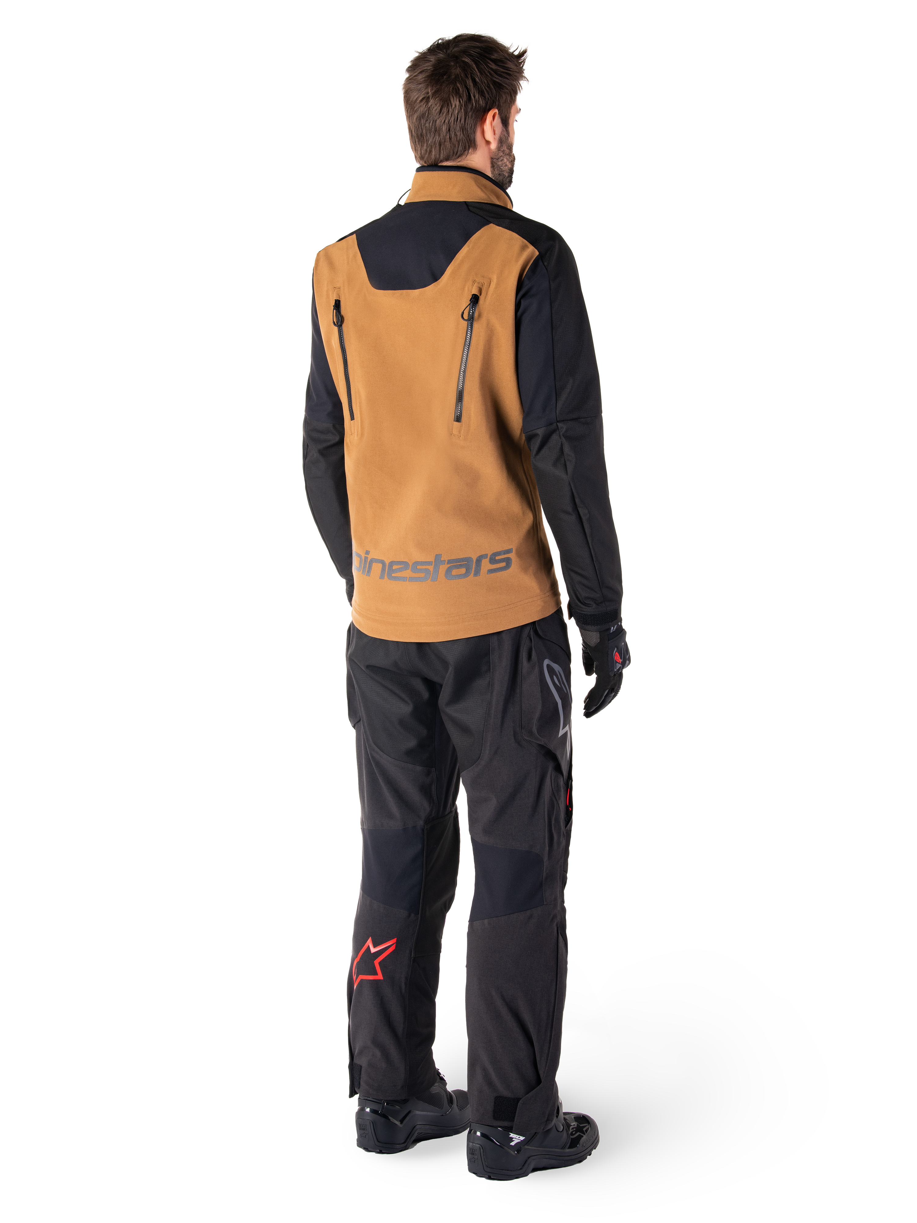 Alpinestars Hyde XT Stretch Drystar®XF-jakke og Hyde XT Drystar®-bukser, set bagfra af en kører iført tan og sort adventure touring-udstyr, jakke med lodrette lynlåse på ryggen og Alpinestars-branding, sorte bukser med rødt stjernelogo på læggen, kombineret med sorte handsker og støvler.
