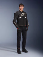 Alpinestars T-GP R V3 Drystar® Jacket in Black and White, worn by a model with black motorcycle gloves and pants, featuring a white chest logo, white sleeve accents, and protective shoulder sliders, waterproof textile sport jacket for road riding