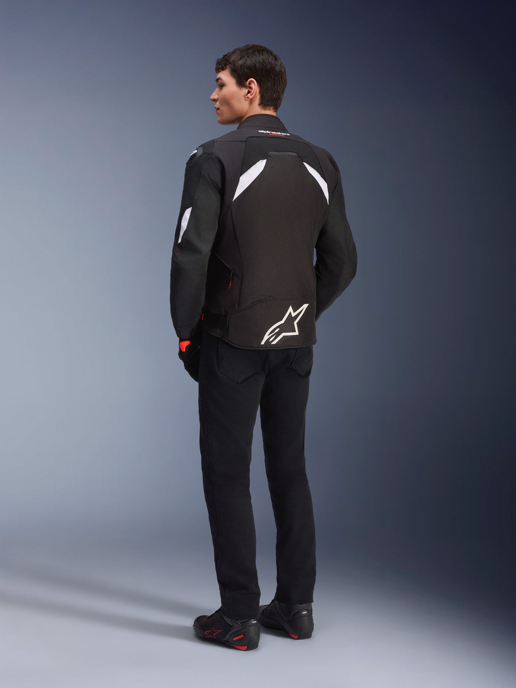 Male model from the back wearing the Alpinestars T-GP R V3 Drystar® Jacket in Black and White, featuring white shoulder accents and a large white logo on the lower back, paired with black riding pants and boots, sport textile motorcycle jacket.