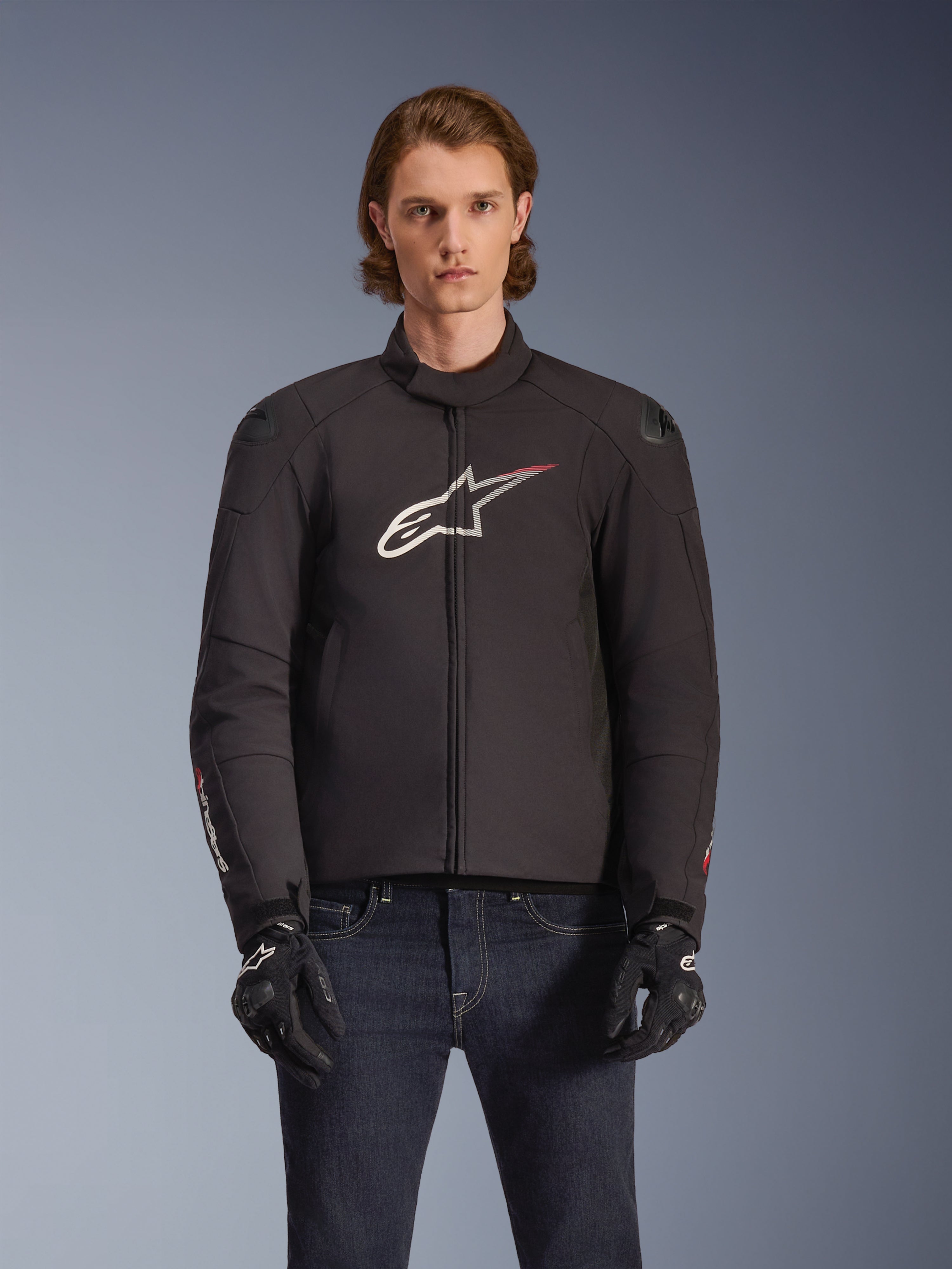 Alpinestars SMX Waterproof Jacket, sort tekstil-motorcykeljakke med hvidt og rødt stjernelogo på brystet, båret af en model med sorte handsker og blå jeans, med udvendige skulder-sliders og en sporty pasform.