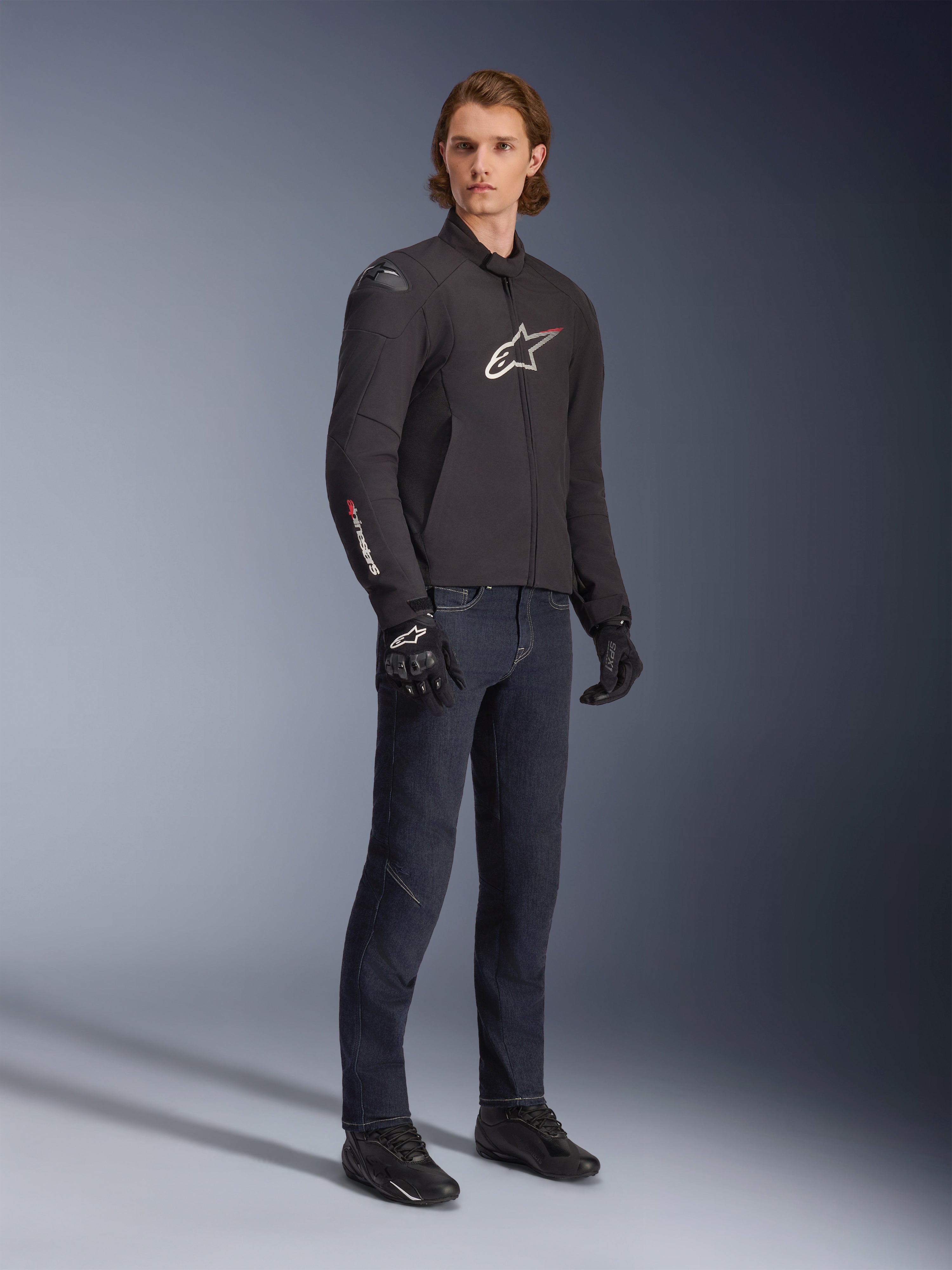Mandlig model iført Alpinestars SMX vandtæt jakke, sort med hvide og røde logo-detaljer, stylet med mørkeblå denimbukser og sorte kørehandsker, strømlinet sport-fit tekstil-motorcykeljakke