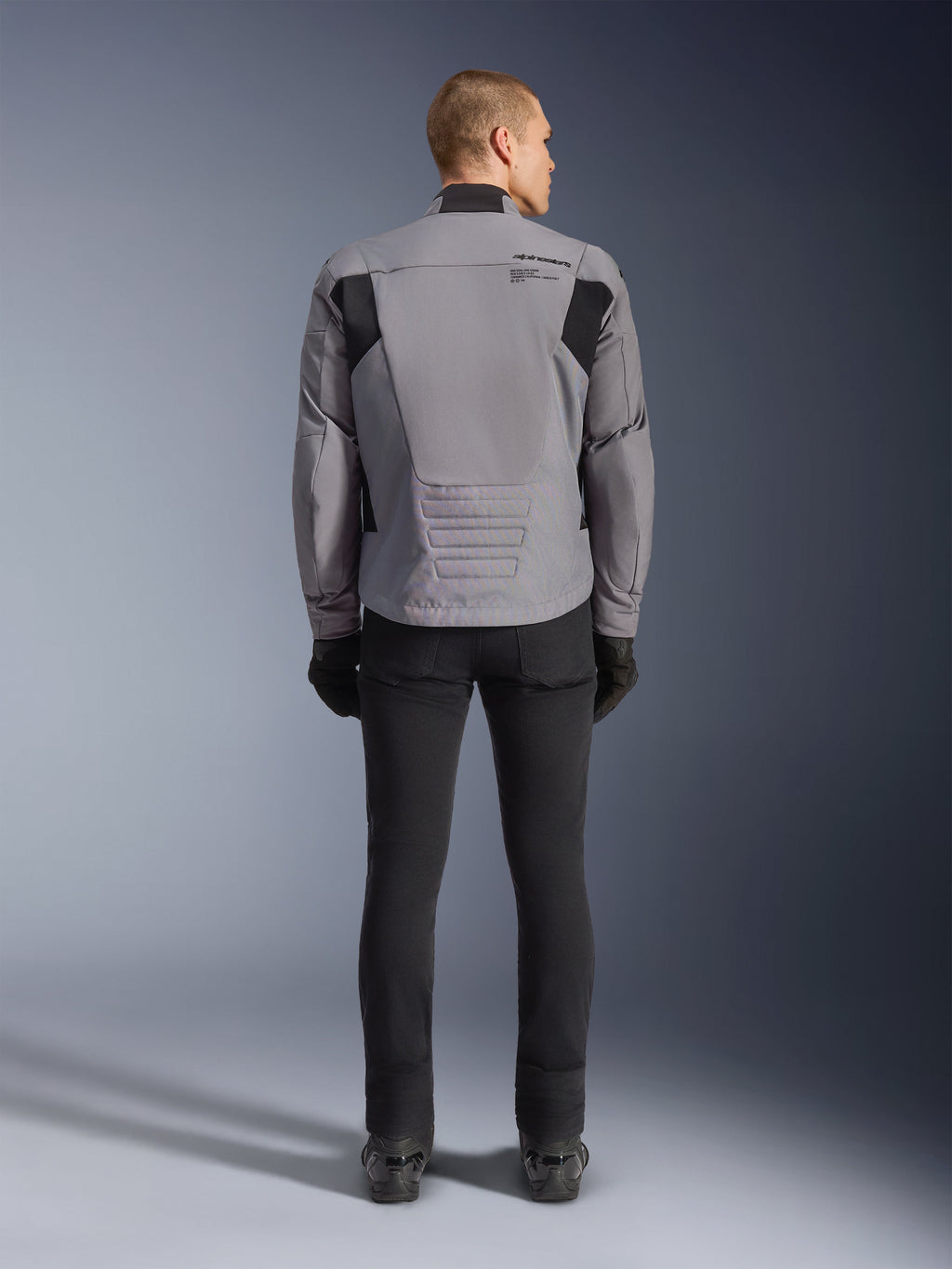 Alpinestars SMX Waterproof jakke, set bagfra, mørkegrå og sort, med sorte tekstilpaneler på skuldrene og et sport-fit design, kombineret med sorte bukser og handsker for et komplet motorcykel-look