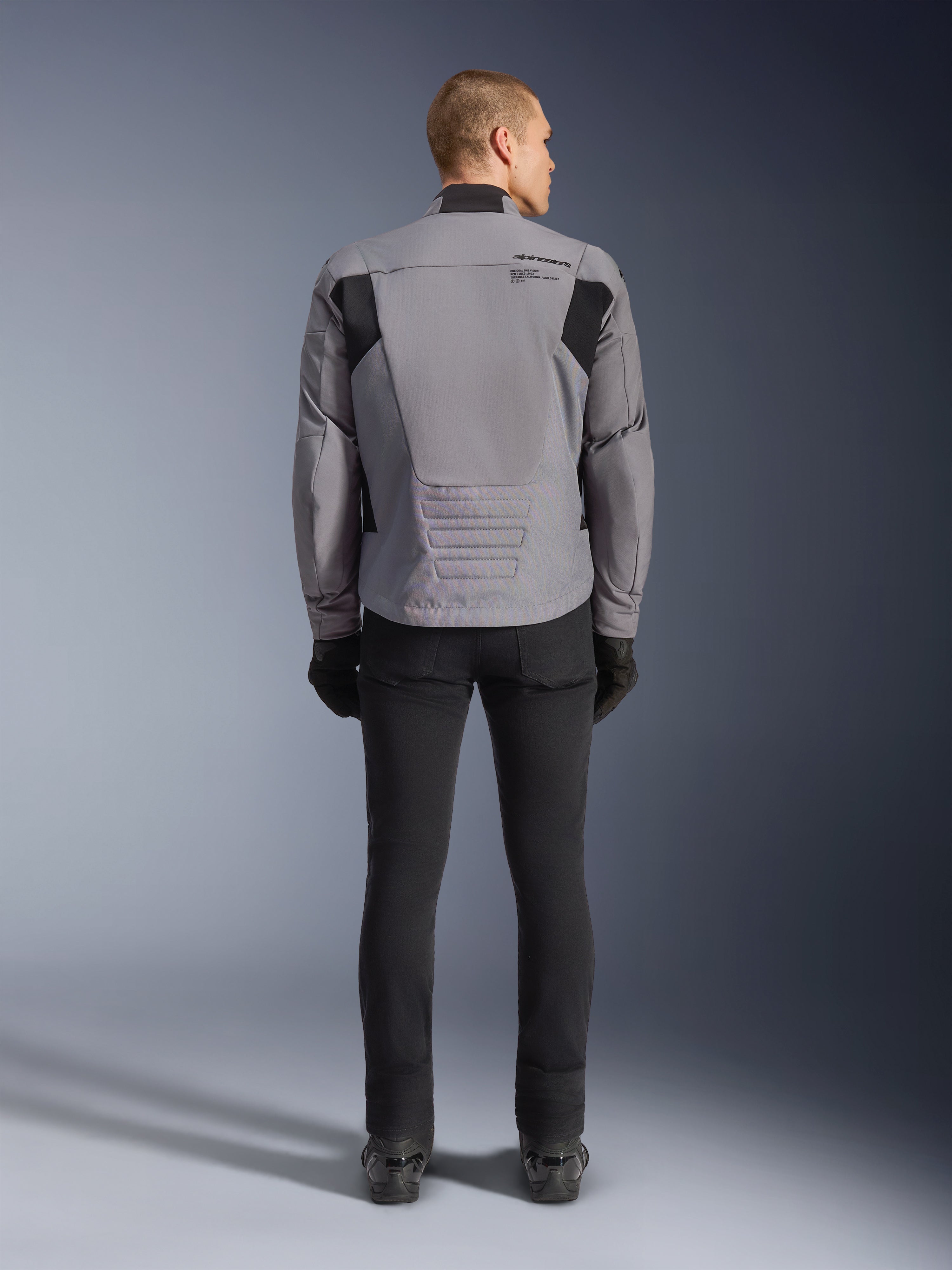 Alpinestars SMX Waterproof jakke, set bagfra, mørkegrå og sort, med sorte tekstilpaneler på skuldrene og et sport-fit design, kombineret med sorte bukser og handsker for et komplet motorcykel-look