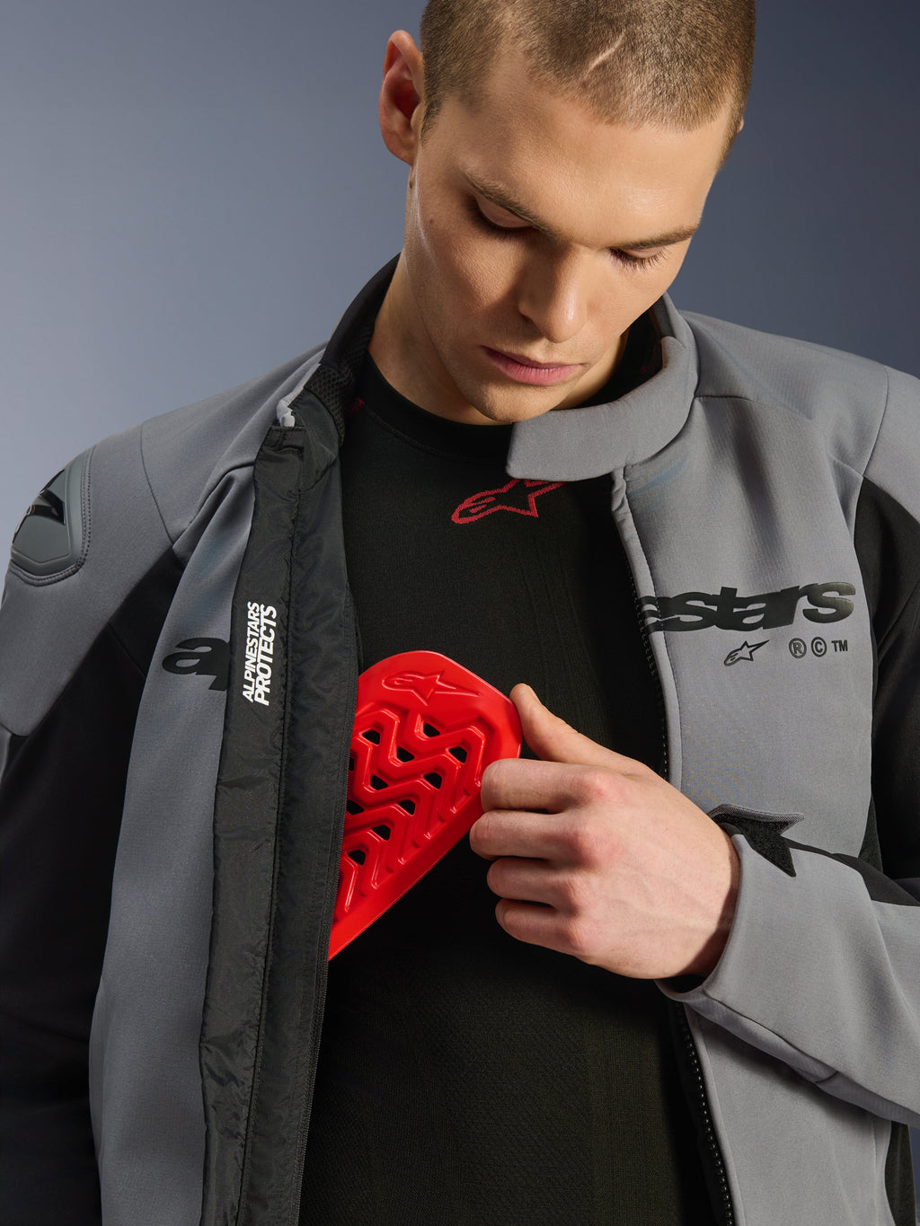 Mandlig model iført Alpinestars SMX vandtæt jakke i mørkegrå og sort, der indsætter en rød Nucleon brystbeskytter i en indvendig lomme for at vise jakkens kompatibilitet med beskyttelse og sikkerhedsfunktioner.