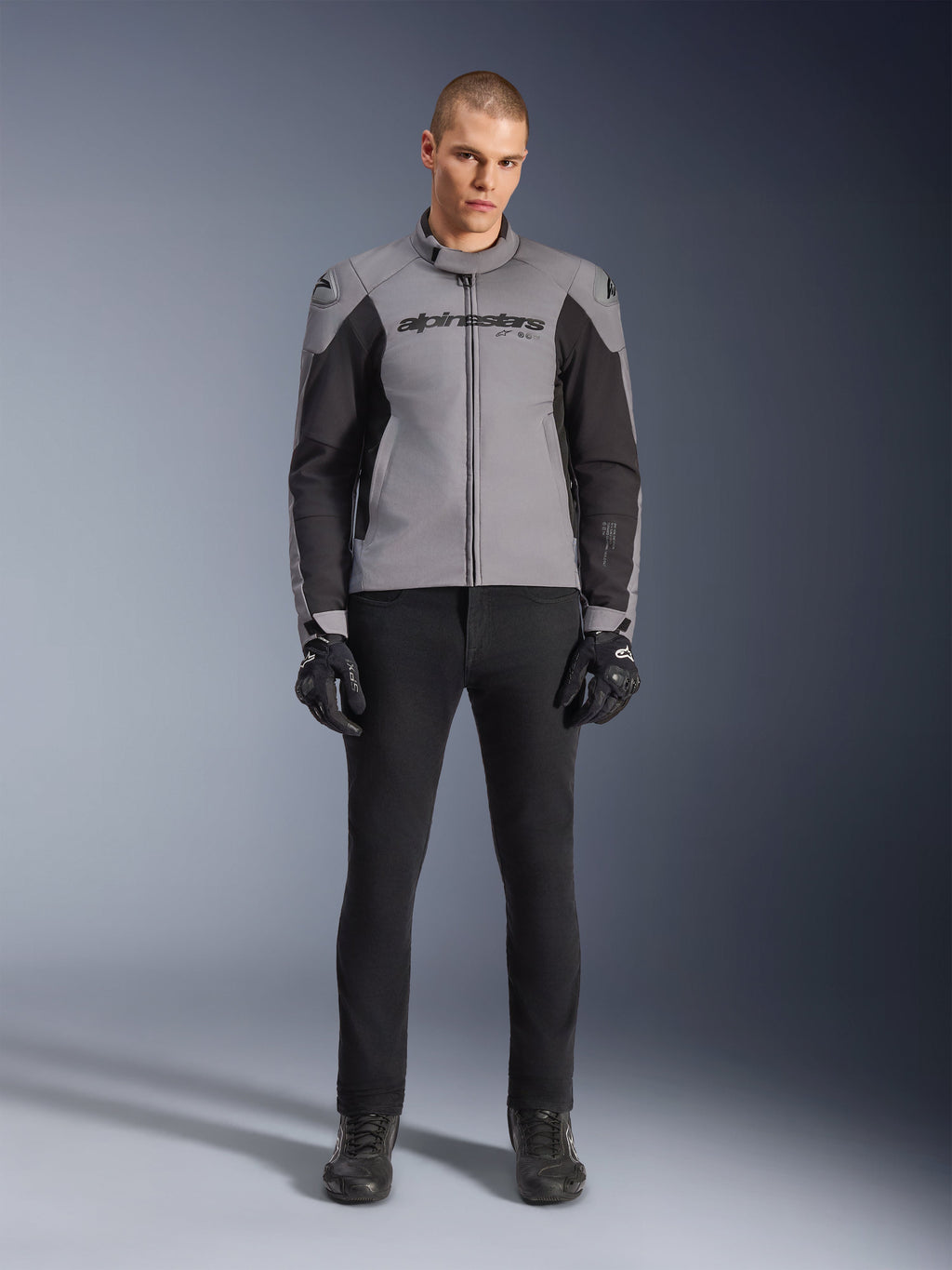 Alpinestars SMX Waterproof jakke, mørkegrå og sort, set forfra på en mandlig model, tekstil sportsmotorcykeljakke med sorte sidepaneler og Alpinestars-branding på brystet, kombineret med sorte kørebukser og handsker