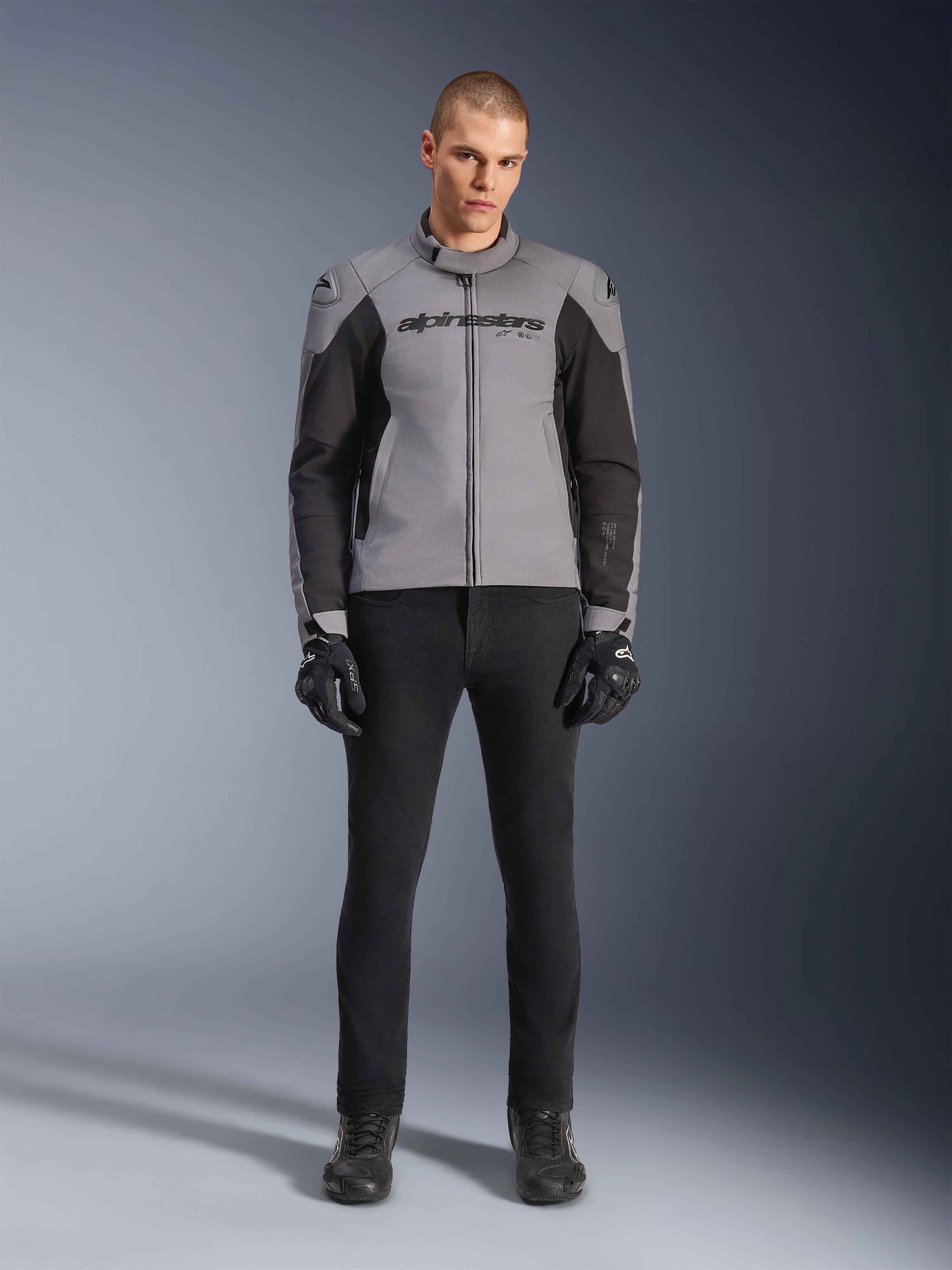 Alpinestars SMX Waterproof jakke, mørkegrå og sort, set forfra på en mandlig model, tekstil sportsmotorcykeljakke med sorte sidepaneler og Alpinestars-branding på brystet, kombineret med sorte kørebukser og handsker
