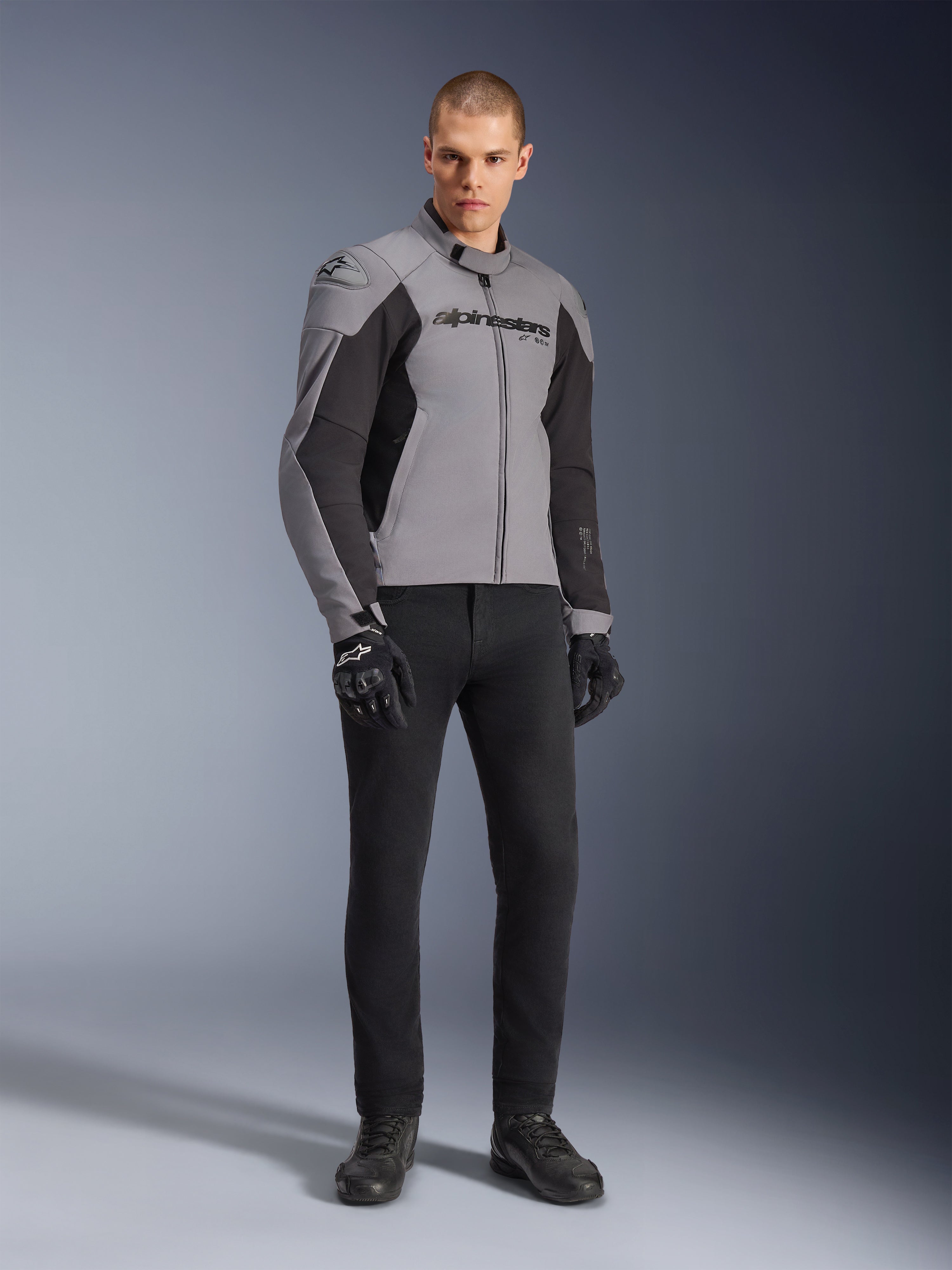 Alpinestars SMX vandtæt jakke, motorcykeljakke, mørkegrå og sort, båret af model med sorte kørehandsker og sorte bukser, med tydeligt logo på brystet og et teknisk sport-mappet design til landevejskørsel i al slags vejr.