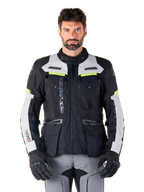 Mand iført Alpinestars Bogotá Pro Drystar-jakke, sort og lysegrå med neongule detaljer, helårs adventure touring motorcykeljakke, vandtæt design med flere lommer og lodret branding, kombineret med sorte handsker og grå kørebukser