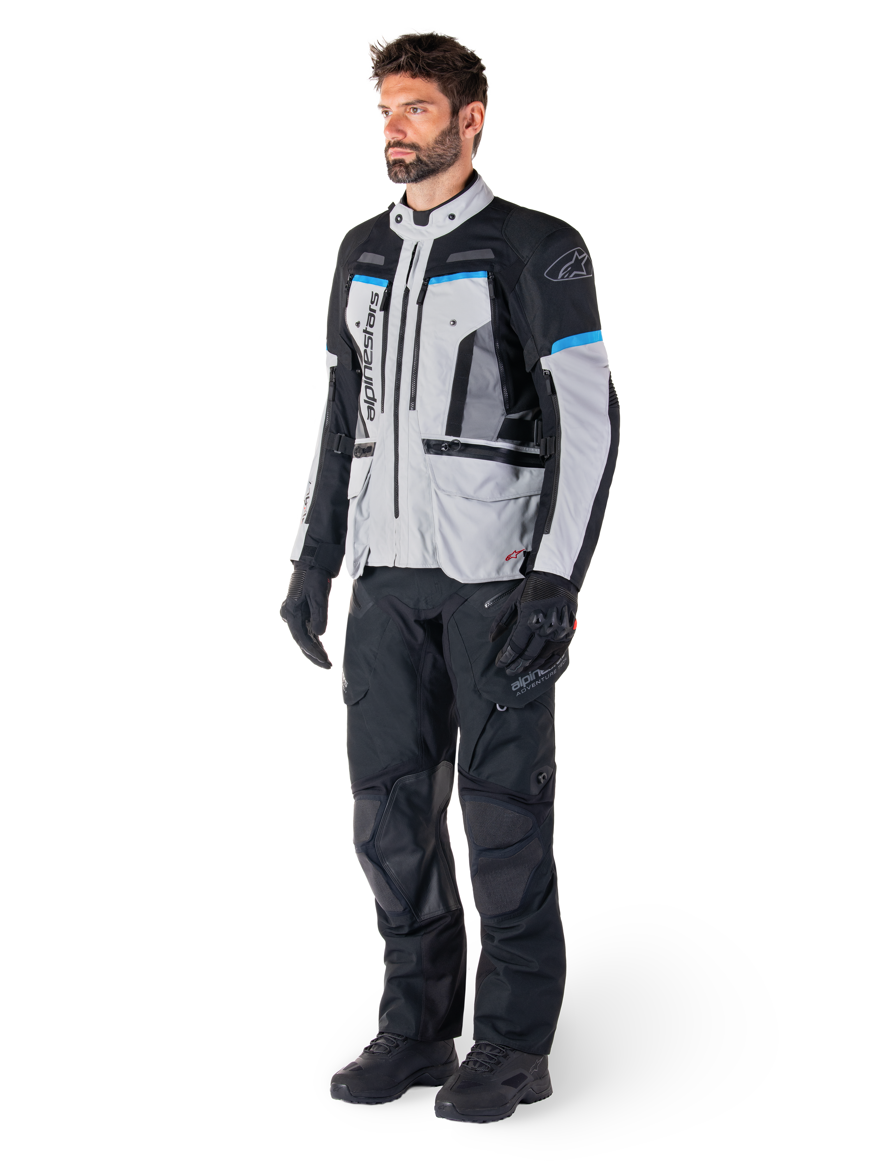 Mandlig kører iført Alpinestars Bogotá Pro Drystar®-jakke i Ice Gray, sort og cyan, kombineret med sorte tekniske kørebukser og handsker, adventure touring-udstyr til ydeevne i al slags vejr.