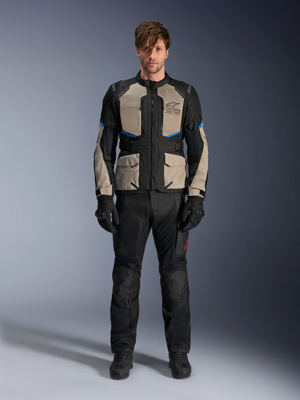 Model iført Alpinestars Andes Air Drystar® jakke, motorcykel touring-jakke, sort, Laurel Gray og lyseblå, kombineret med matchende sorte kørebukser og handsker, med omfattende mesh-ventilationspaneler på brystet og ærmerne til adventure touring i varmt vejr.