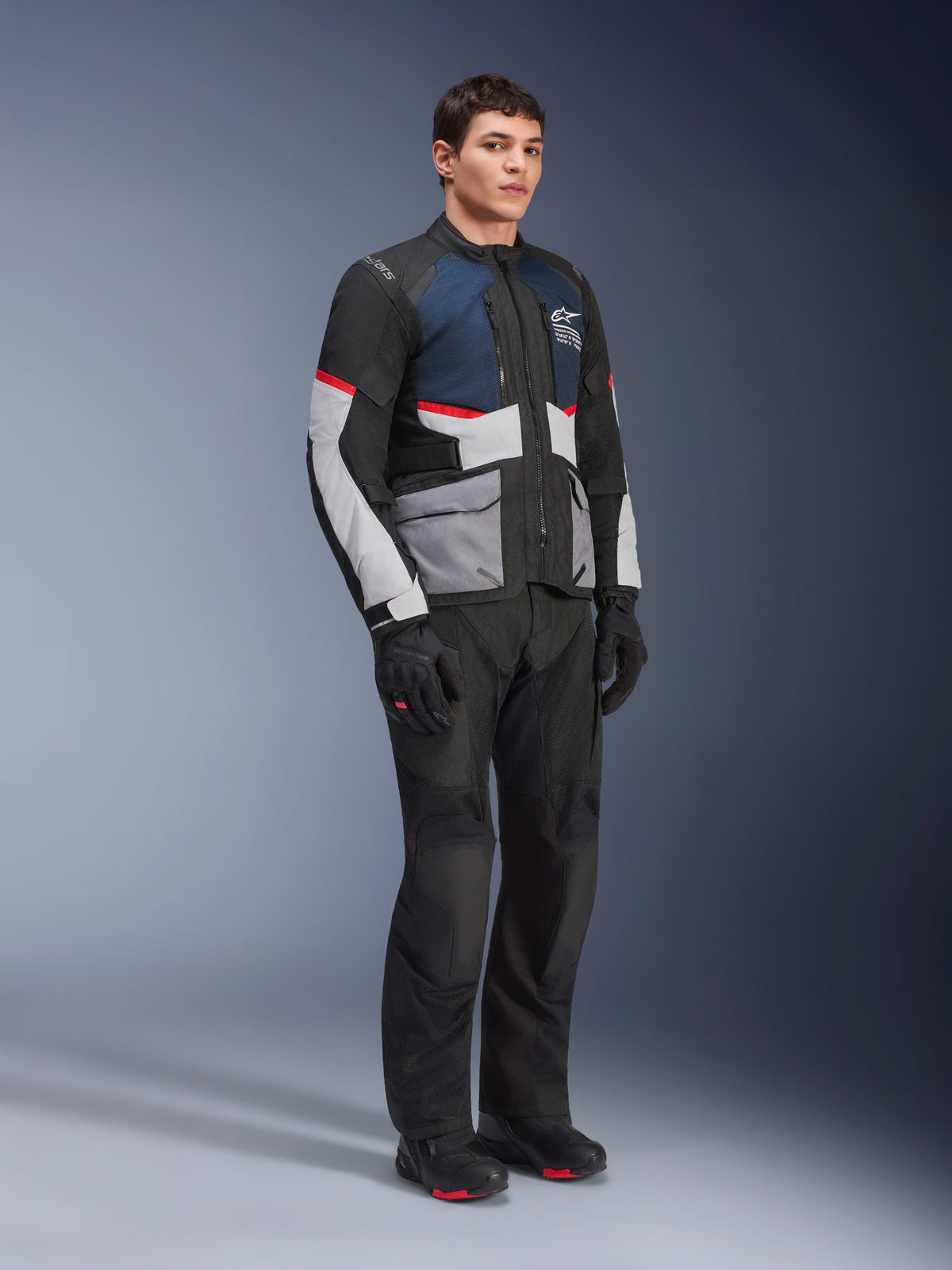 Mandlig model iført Alpinestars Andes Air Drystar® jakke i Deep Blue Black Ice Gray, kombineret med sorte motorcykelbukser, handsker og støvler, med store mesh-paneler til ventilation, lysegrå lommer og røde detaljer, designet til adventure touring.