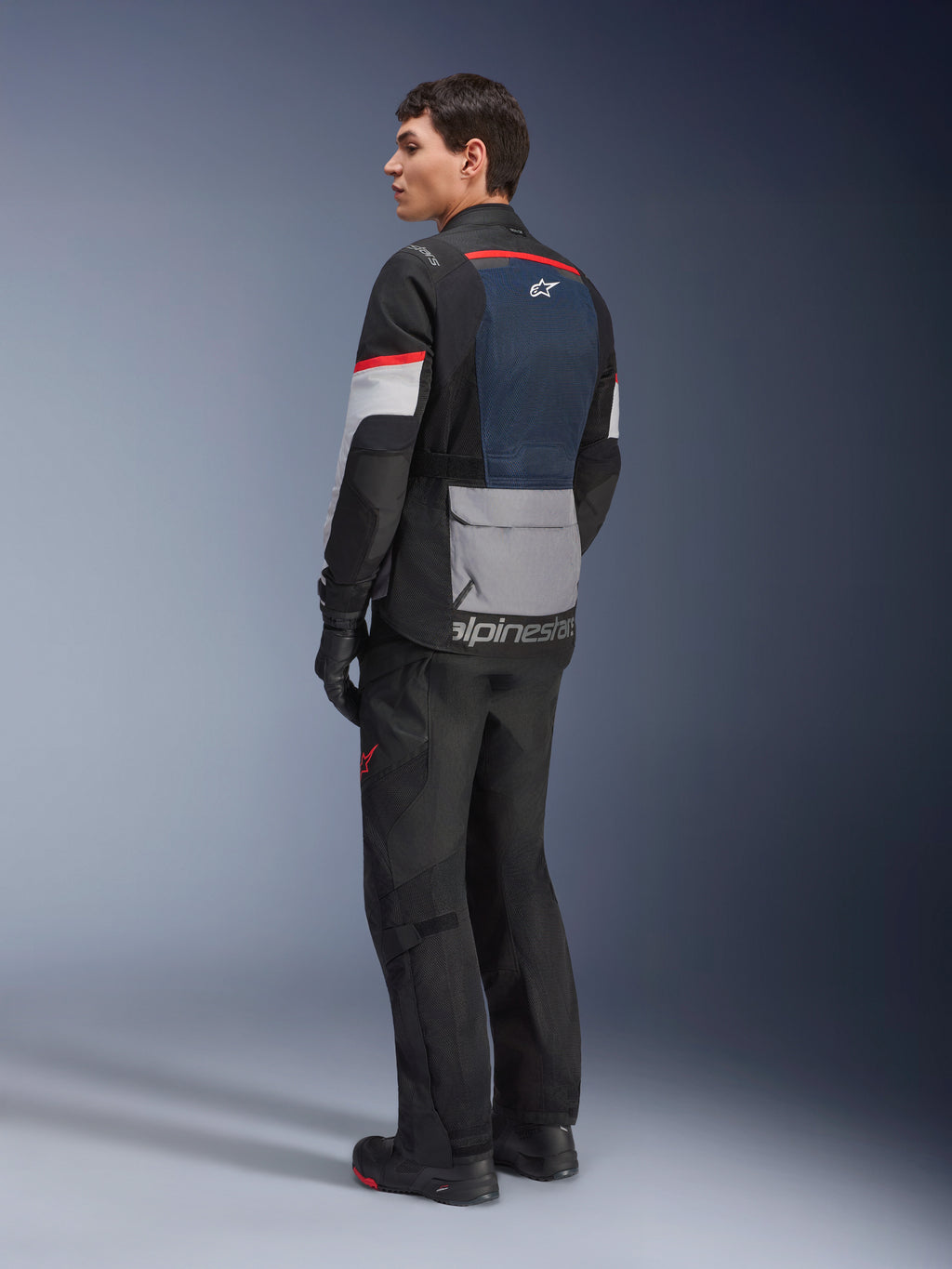 Mandlig model set skråt bagfra iført Alpinestars Andes Air Drystar®-jakke i Deep Blue Black Ice Gray, kombineret med sorte motorcykelbukser og handsker, med blåt mesh-rygpanel, grå utility-lomme på lænden og røde detaljer på ærmerne, professionelt adventure touring-udstyr.
