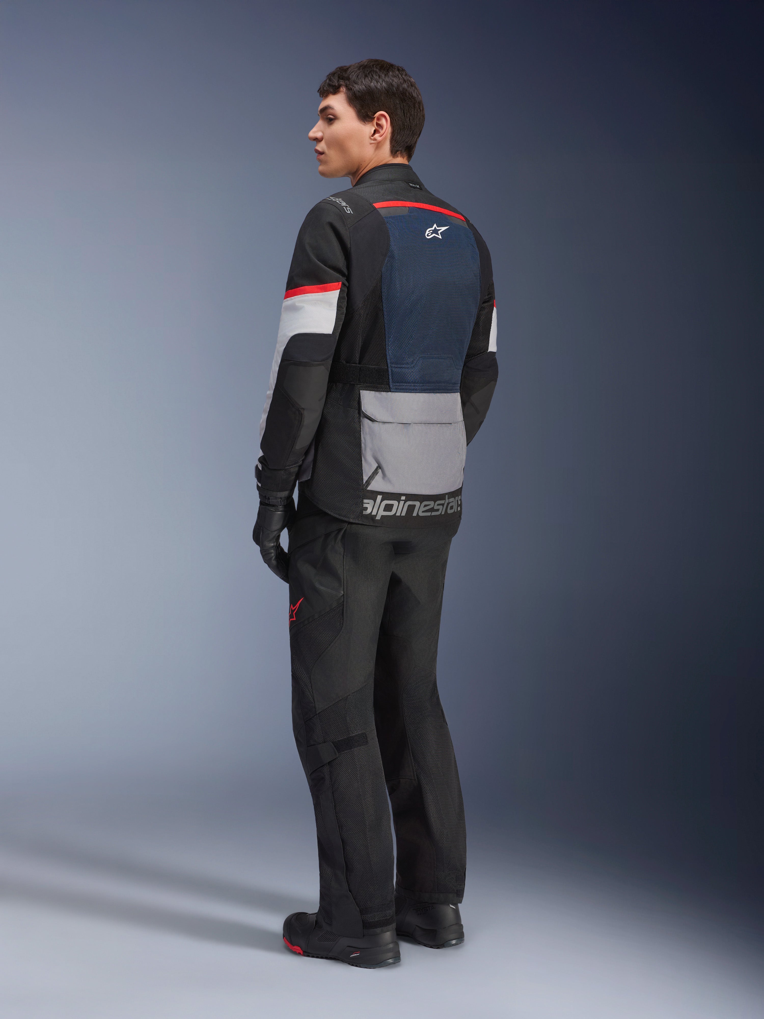 Mandlig model set skråt bagfra iført Alpinestars Andes Air Drystar®-jakke i Deep Blue Black Ice Gray, kombineret med sorte motorcykelbukser og handsker, med blåt mesh-rygpanel, grå utility-lomme på lænden og røde detaljer på ærmerne, professionelt adventure touring-udstyr.